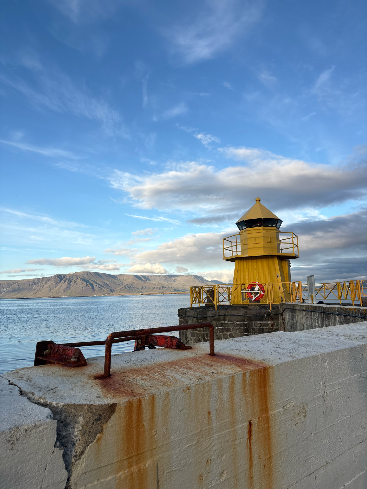 A bright yellow lighthouse stands on a stone pier overlooking a calm body of water. In the background, a range of mountains stretches across the horizon under a partially cloudy sky. The lighthouse has a red life preserver attached to its exterior. The foreground features a weathered concrete pier with visible rust stains and a large crack. A railing made of rusty metal is also present on the pier. The scene appears to be a harbor or coastal area in Reykjavík, Iceland.