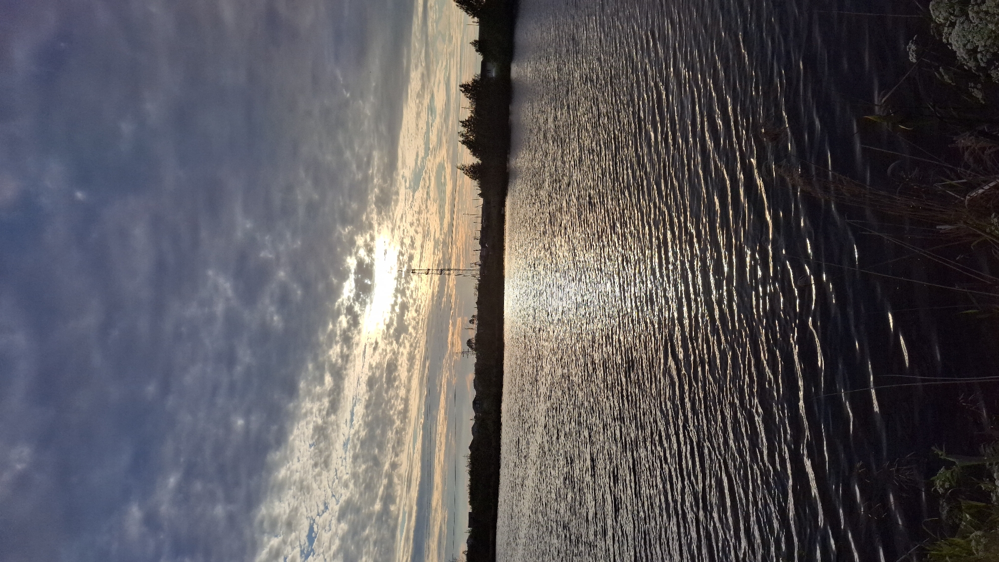This is a vertical, eye-level shot of a body of water in Innolovo, Russia, at dusk. The water's surface is rippled, reflecting the light of the setting sun, which is partially obscured by clouds. A power line tower stands on the horizon, silhouetted against the sky. The shoreline is lined with trees and what appear to be buildings. The foreground is dominated by the dark water, with some vegetation visible at the bottom of the frame. The sky is a mix of dark gray clouds and lighter, scattered clouds, with a bright orange glow emanating from the sun.