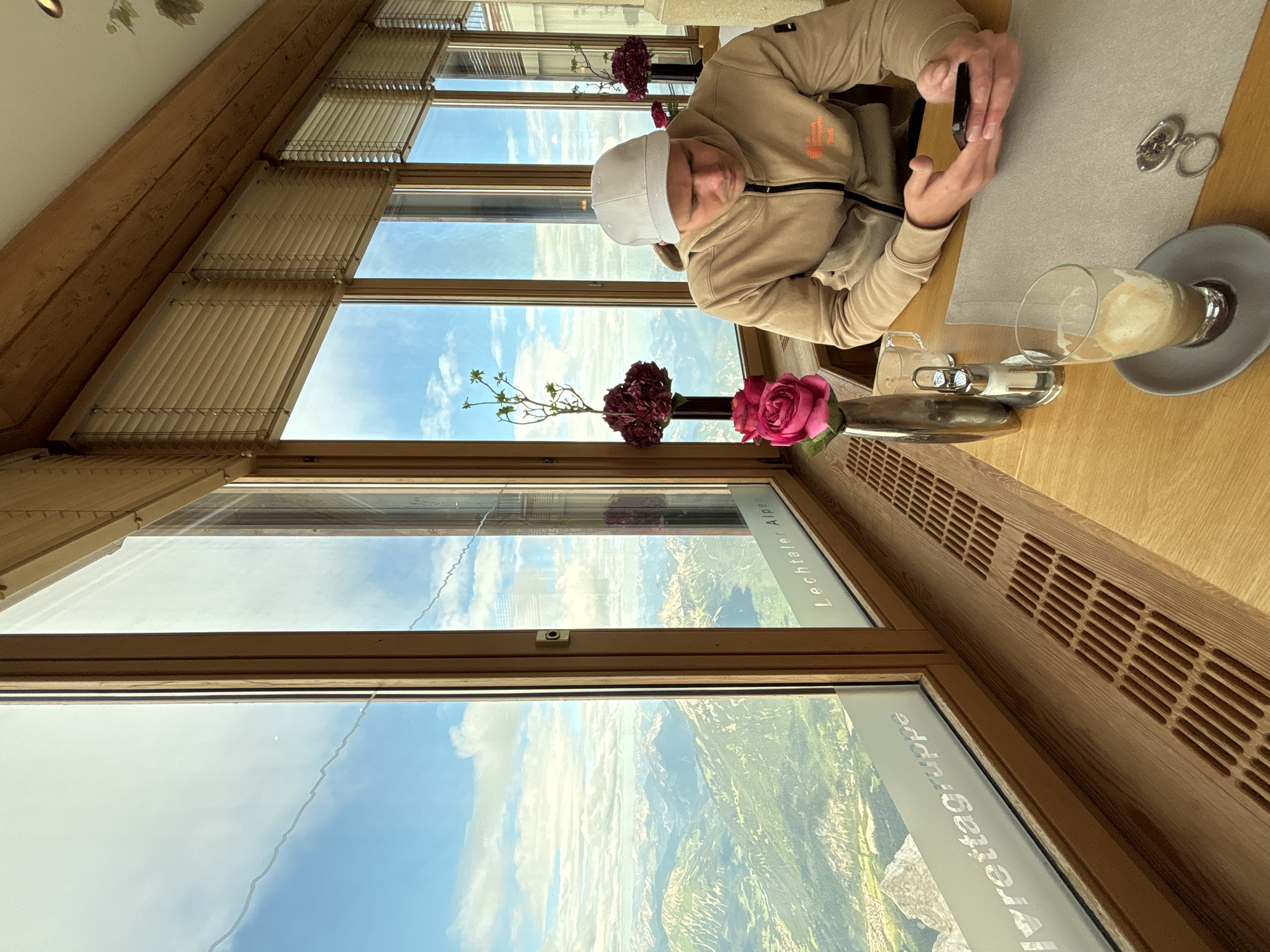 A man wearing a light-colored baseball cap and a tan hooded sweatshirt is seated at a wooden table, looking at his smartphone. He is positioned in the foreground, to the right of the frame, with a glass of coffee and a salt and pepper shaker on the table in front of him. Behind the man, a large window offers a panoramic view of a mountain range under a partly cloudy sky. The window is framed by wooden beams and blinds. The text "Lechtaler Alpen" and "lvreittagruppe" are visible on the window. The setting appears to be a restaurant or cafe with large windows, likely at a high elevation in the Austrian Alps near Ehrwald, Austria.
