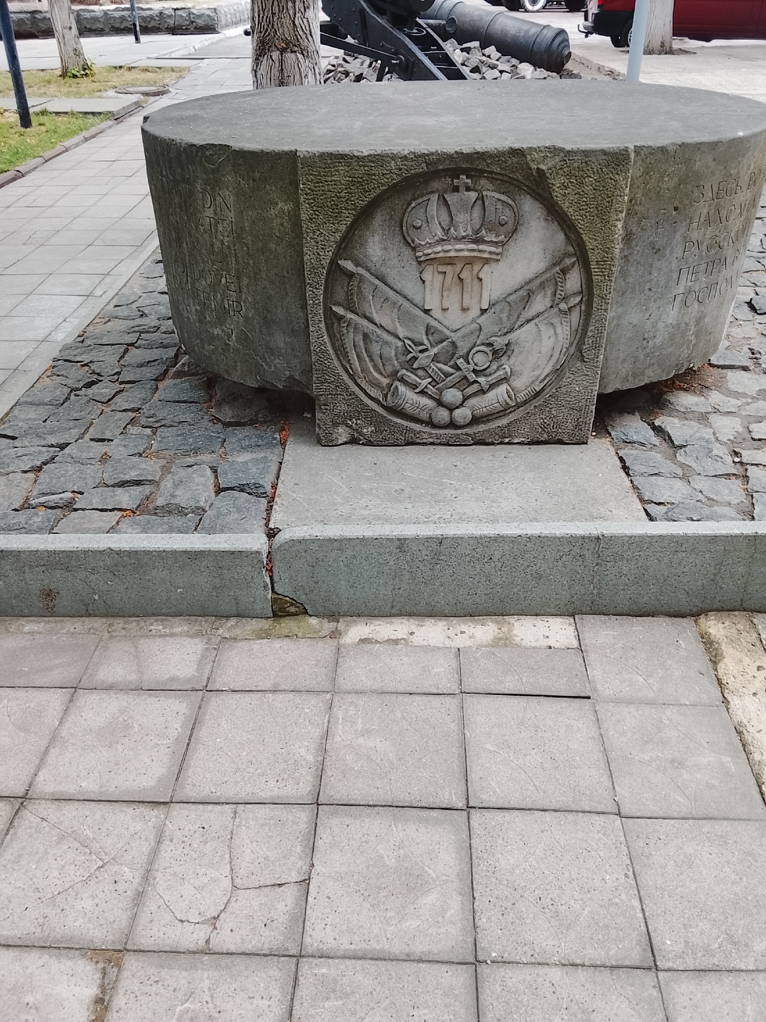The image shows a stone monument on a sidewalk in Chișinău, Moldova. The monument is hexagonal and made of rough stone, with a circular relief carved into its front. The relief depicts a crown, the year "1711", and two crossed swords. To the right of the relief, there is an inscription in Russian that reads "ЗДЕСЬ В 1711 НАХОДИЛАСЬ РУССКАЯ ГОСПИТАЛЬ ПЕТРА I" which translates to "HERE IN 1711 WAS THE RUSSIAN HOSPITAL OF PETER I". The monument is situated on a cobblestone base, which is then on a concrete platform, and finally on a tiled sidewalk. In the background, a historical cannon and trees are visible, suggesting a park or historical site.