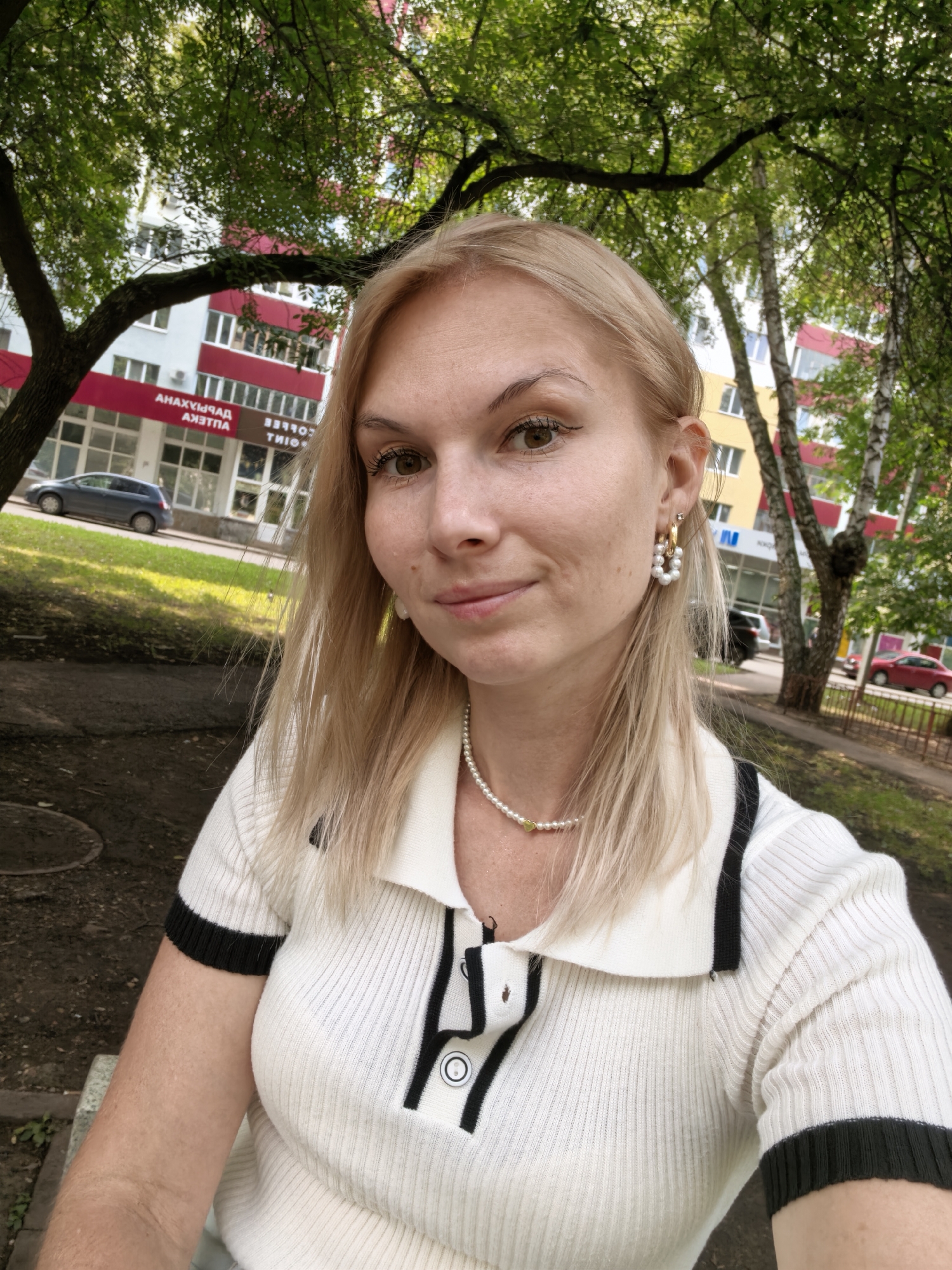 A young Caucasian woman with blonde hair and brown eyes is captured in a selfie outdoors. She is wearing a white collared shirt with black trim and buttons, and a pearl necklace with a small gold heart pendant. She has on gold hoop earrings with pearls. The background shows a residential building with red and yellow accents, green trees, and a street with cars. The scene appears to be in a park or public space in the city of Sterlitamak, Russia, given the presence of the building with Cyrillic signage that reads "АНАХЫМЫЯД АЗЯТПА". The lighting suggests it is daytime.