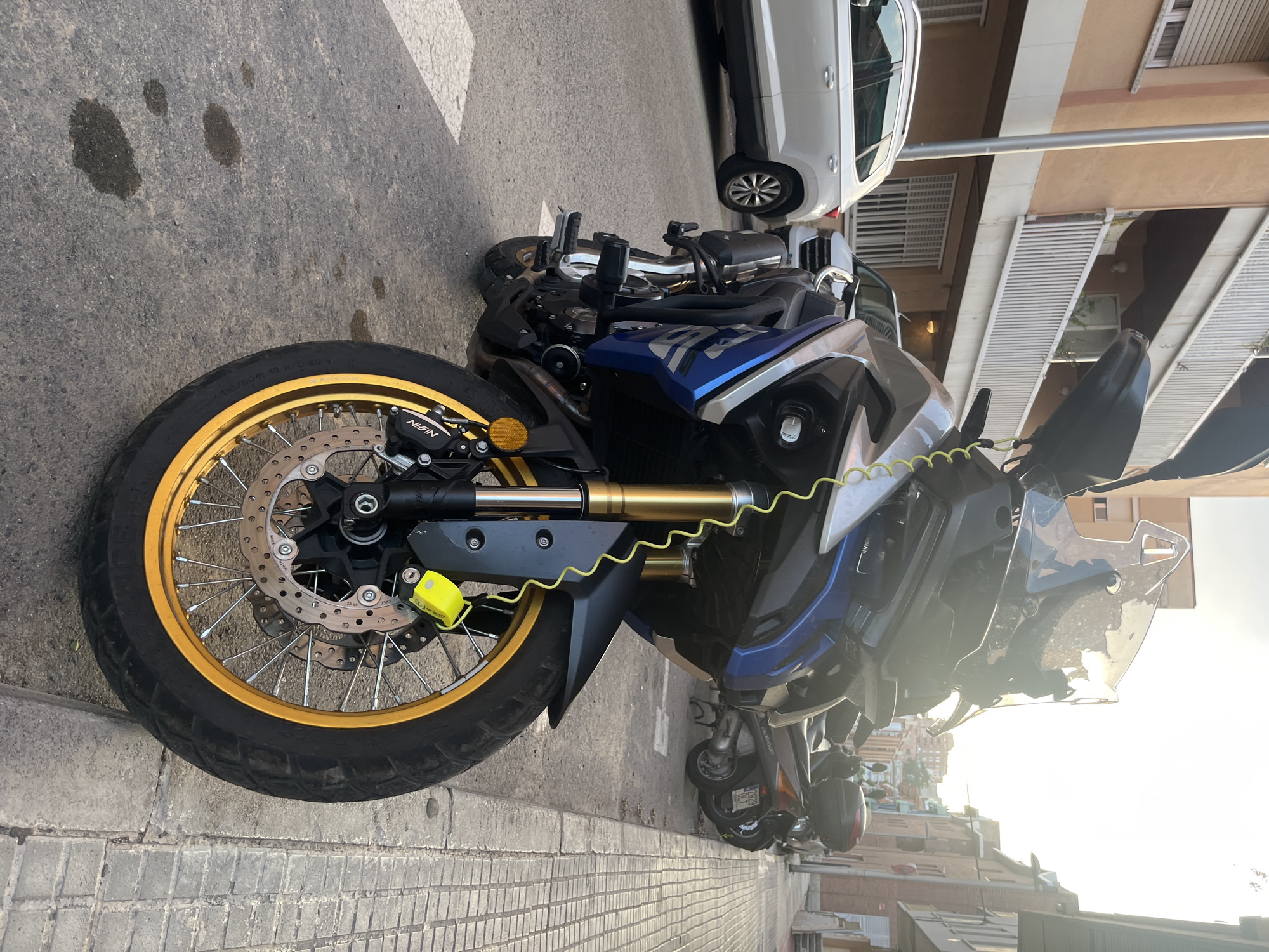 The image depicts a blue and silver motorcycle parked on an asphalt street in Tarragona, Spain, with a yellow security cable looped through its front brake disc and attached to a bright yellow lock. The motorcycle is positioned in the foreground, facing slightly towards the viewer's left. Its front wheel, with a gold rim and spokes, is clearly visible, along with part of the suspension and braking system.
Behind the motorcycle, several scooters are parked along the curb. To the left of the motorcycle, a white SUV is parked. The background features buildings with balconies and windows, characteristic of urban architecture in Spain. The lighting suggests it is late afternoon or early evening, with the sun casting long shadows and creating a warm glow. The street appears to be a typical urban environment, with a sidewalk made of concrete tiles. There are several dark, circular stains on the asphalt in front of the motorcycle, possibly oil or water. No people are visible in the image.