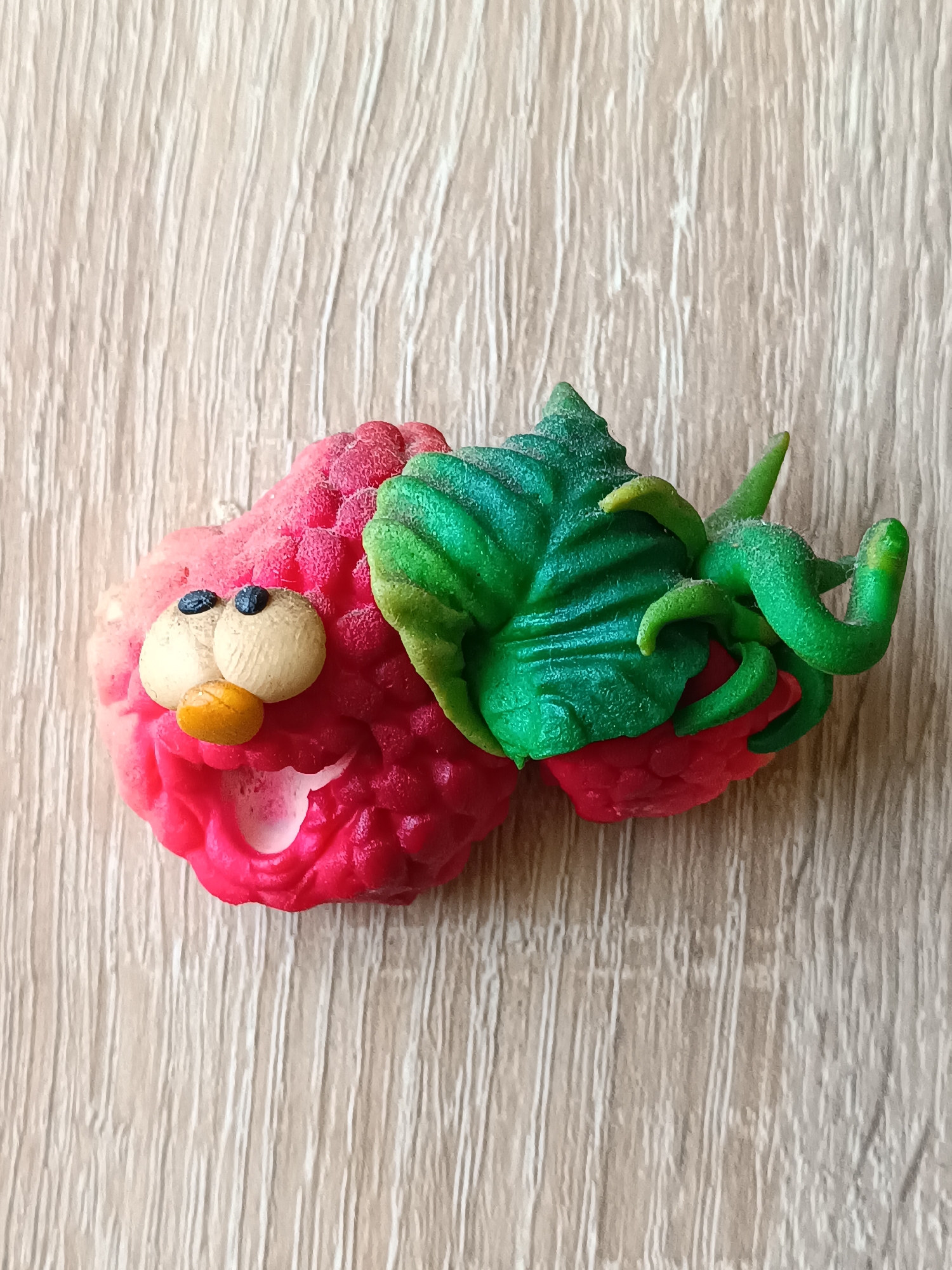 This is a top-down, medium shot photograph of a whimsical, handmade raspberry character crafted from modeling clay. The character is positioned on a light-colored wooden surface with a visible grain pattern. The raspberry character features a bright pink and red textured body, mimicking the drupelets of a real raspberry. It has two circular, light-colored eyes with black pupils, and a small, bright orange nose. A wide, open mouth with a white interior suggests a smiling or exclamatory expression. Attached to the right side of the raspberry is a vibrant green leaf with textured veins and several curly green stems. The overall impression is playful and cartoon-like. The image does not contain any elements that would indicate a specific location in the city of Tebras, Latvia, or any other geographical context. The scene is static and there are no people or animals present.