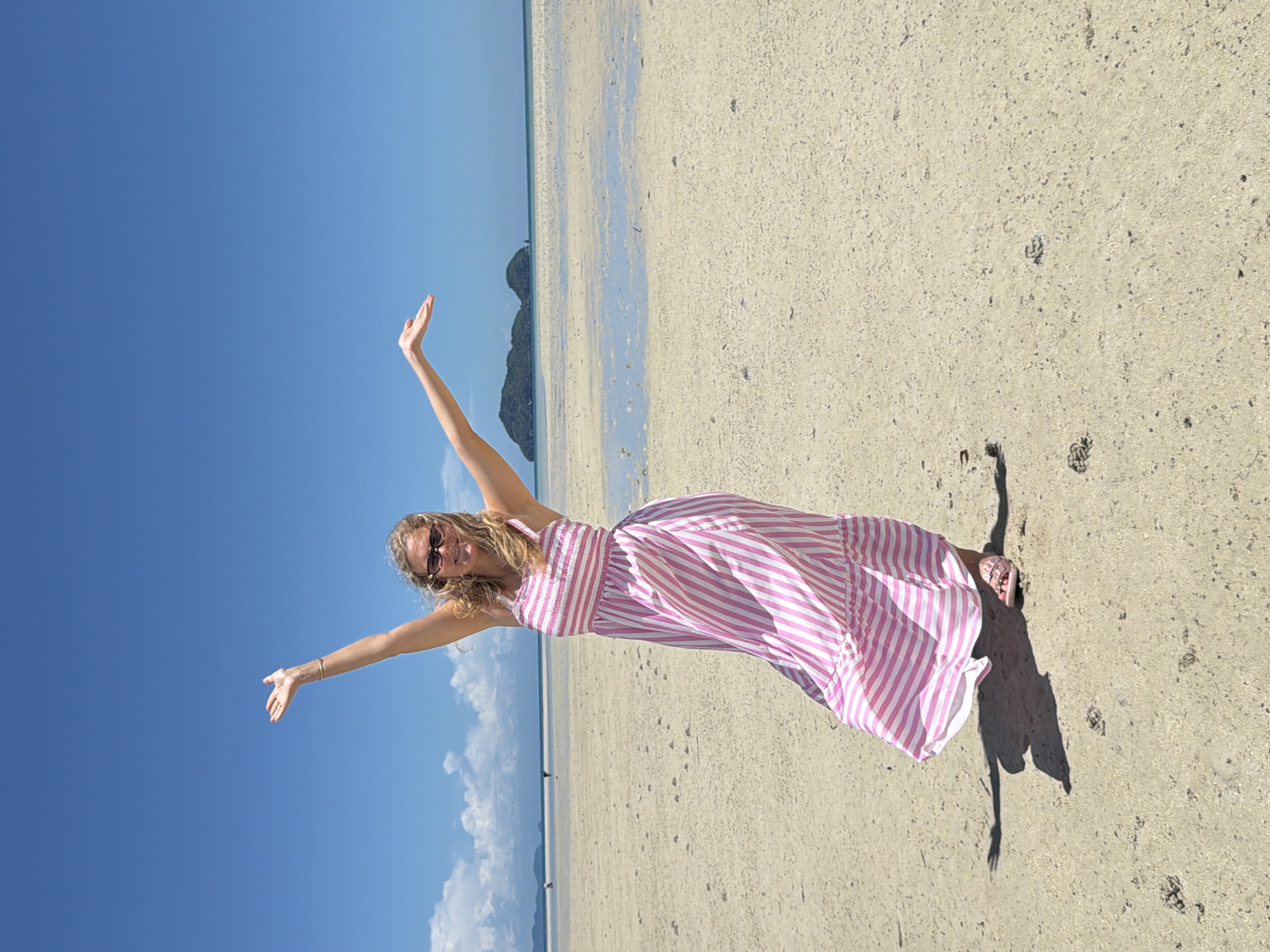 A woman stands on a sandy beach under a clear blue sky, with her arms raised and spread wide in a gesture of joy or welcome. She is wearing a long, pink and white striped dress and sunglasses. The beach is wet in some areas, with shallow pools of water reflecting the sky. In the distance, there are two small, green islands. The sand is dotted with small shells and footprints. The scene appears to be at low tide. The location is identified as Tambon Ko Pha Ngan, Thailand.
