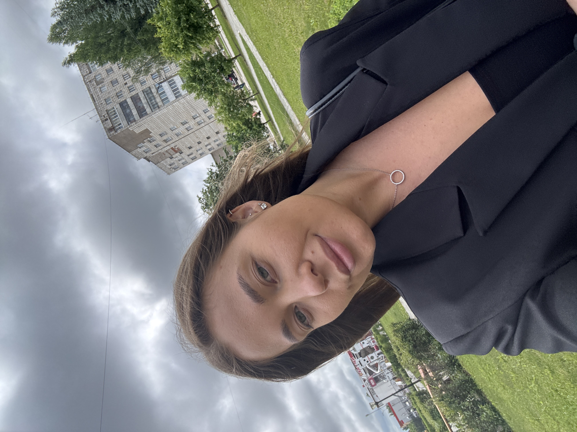 A young woman with light brown hair is taking a selfie outdoors. She is wearing a black blazer over a black top and a silver necklace with a circular pendant. The background shows a cloudy sky, a large residential building, trees, a grassy area, and a building with red accents and a fire truck visible. This scene is located in the city of Chernushka, Russia.