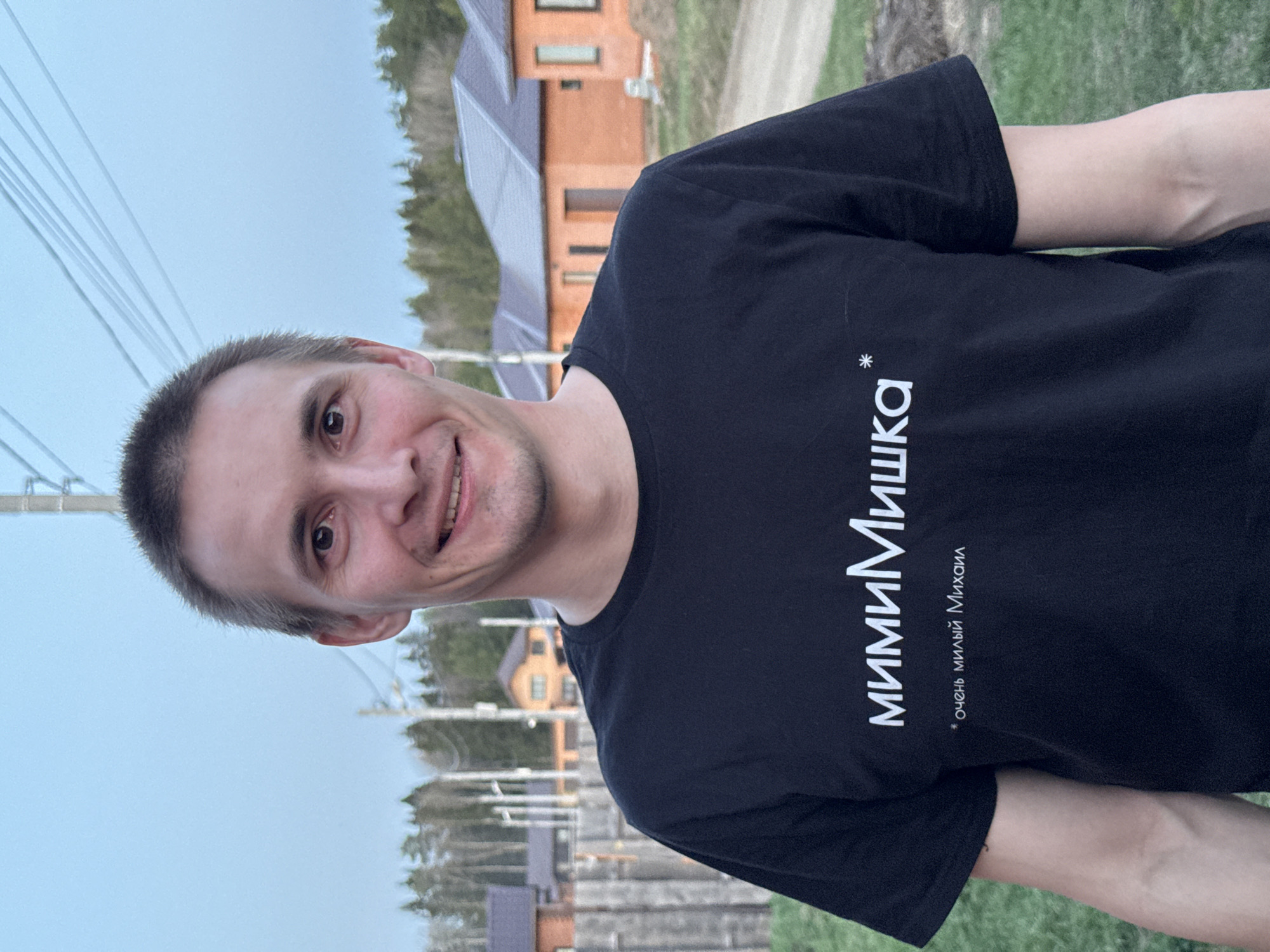 A man is pictured wearing a black t-shirt with white Cyrillic text on it that translates to "Mimi Mishka" and below it "* Very sweet Michael". He is facing forward with a slight smile. The background features a mix of residential buildings, power lines, and greenery. The location appears to be in a residential area of Chaikovsky, Russia.