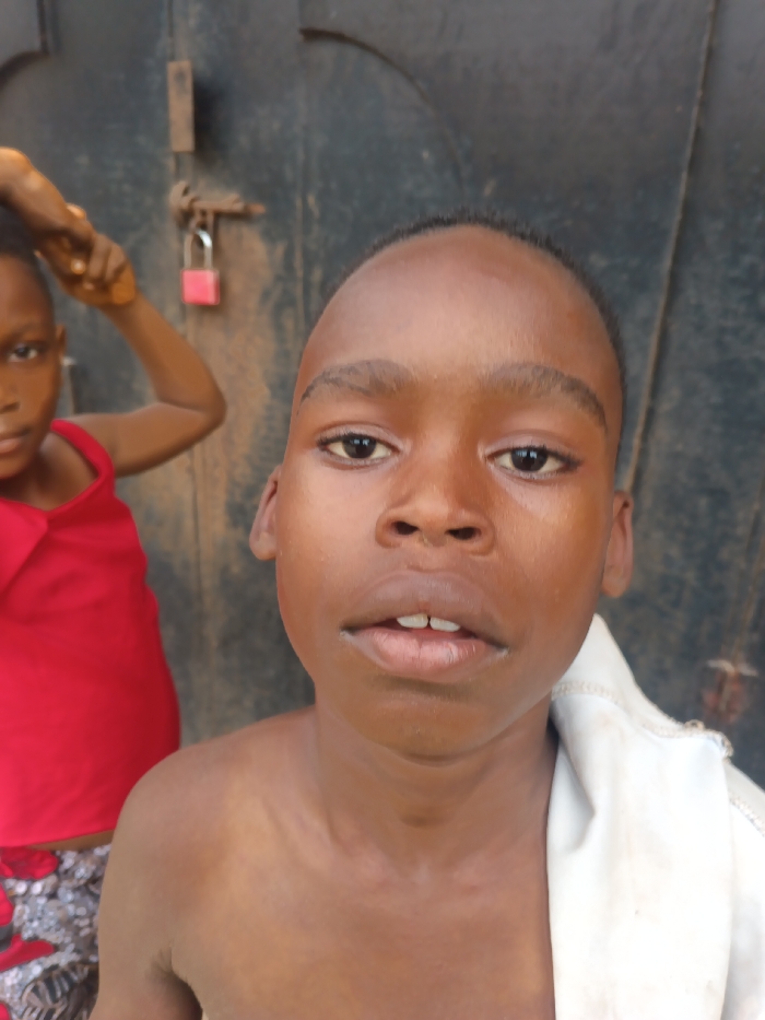 Two young individuals are depicted in Itu, Nigeria, positioned in front of a dark, weathered metal gate secured by a silver latch and a bright pink padlock. In the foreground, a young male with dark skin, short dark hair, and dark eyes looks directly at the camera, his mouth slightly ajar, revealing upper teeth. He is shirtless, with a white fabric garment draped over his left shoulder. Partially obscured and out of focus in the background to the left is a second young individual, also with dark skin, wearing a red sleeveless top. This individual's right arm is raised, with the hand near the head. The visible ground beneath is brownish.