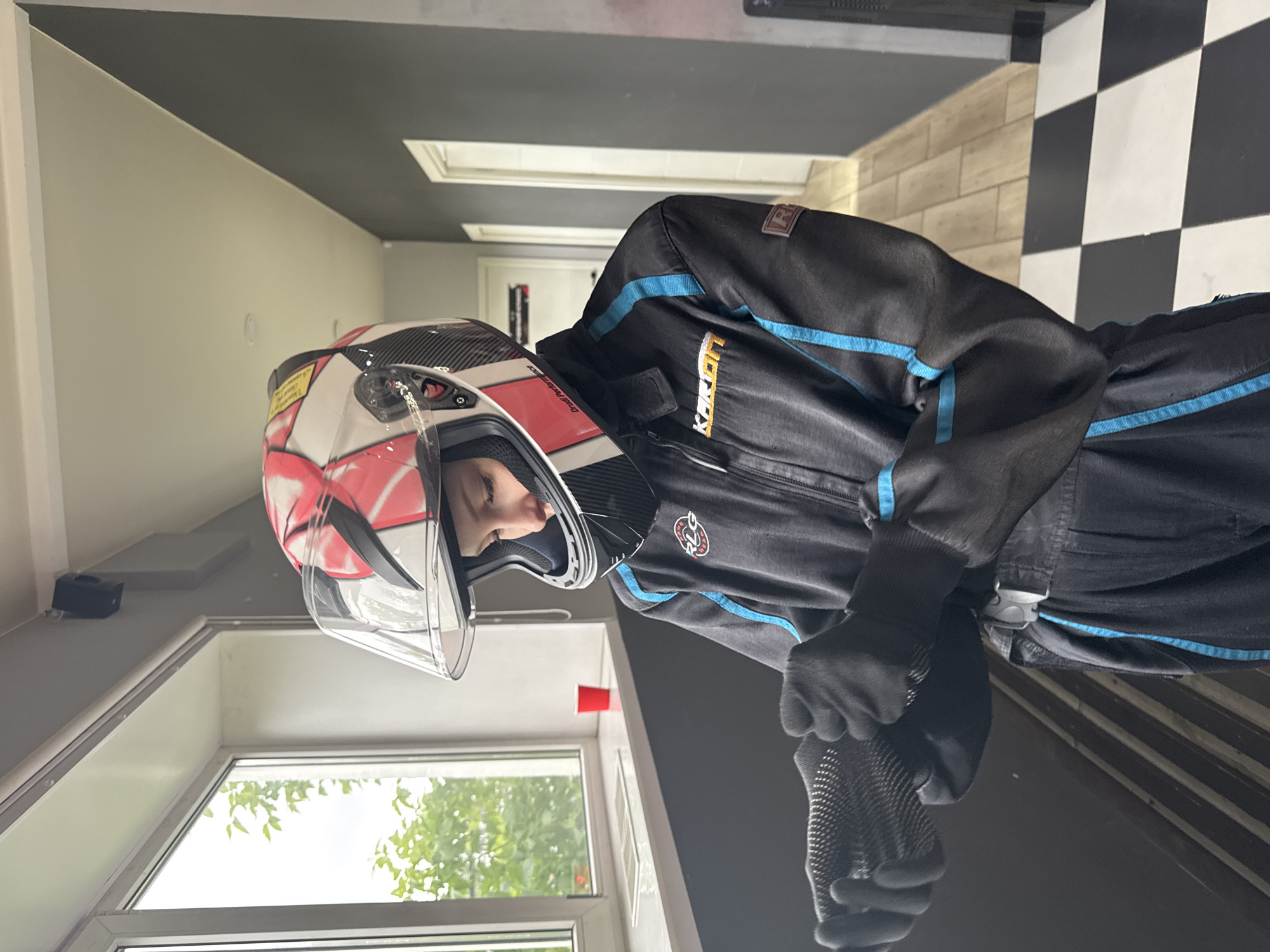 A young person wearing a black go-karting suit with blue trim, black gloves, and a red and white racing helmet with a clear visor is standing indoors. The background features a window with trees visible outside, a checkered floor pattern, and a door. The suit has the logo "Karland" on the upper right side. The location is likely a go-karting center in Perm, Russia. The person is standing still, possibly preparing for or after go-karting.