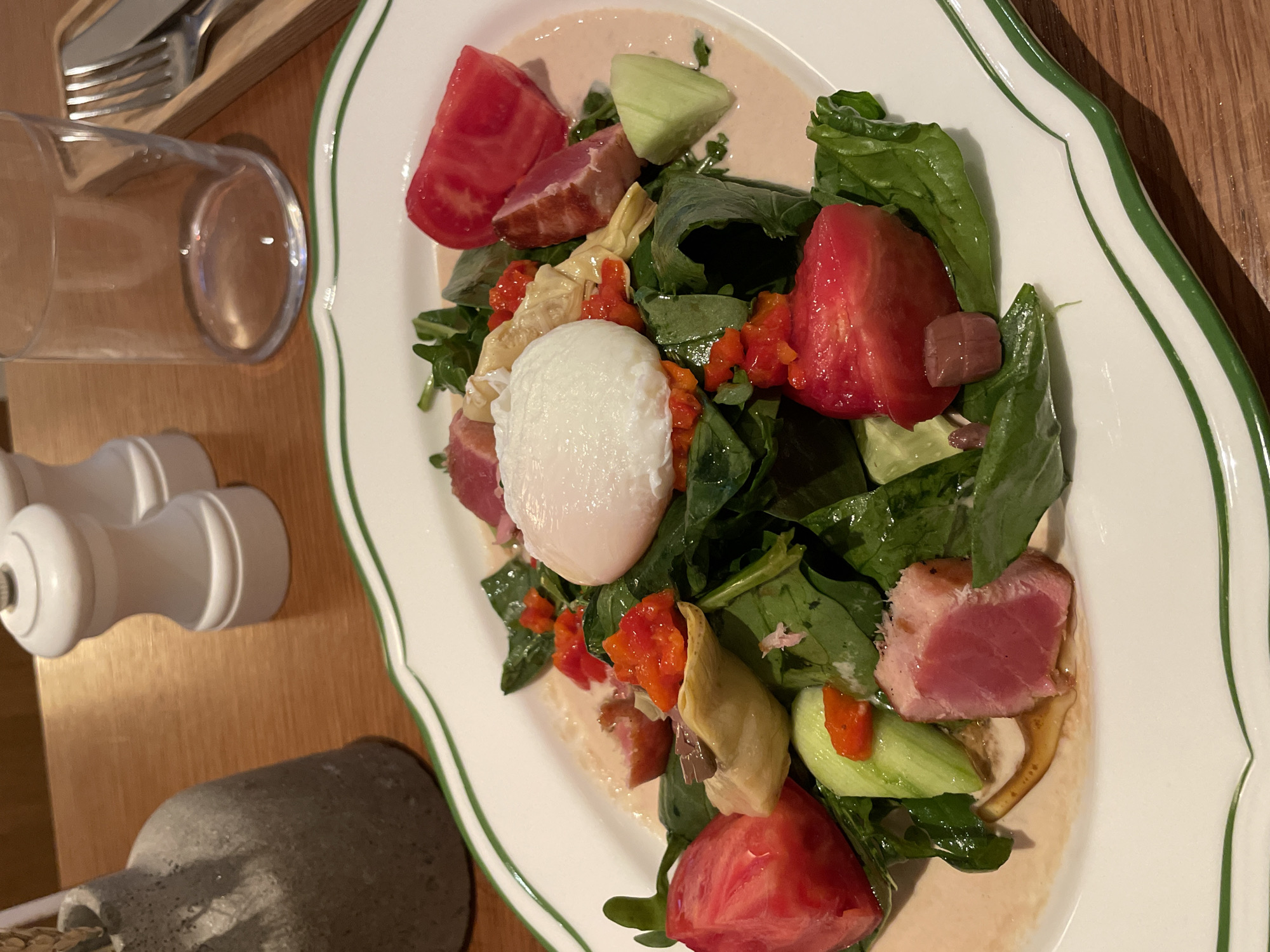 The image shows a plate of salad served on a white plate with a green border. The salad consists of various components, including green leafy vegetables, chunks of red tomatoes, cucumber slices, pieces of cooked tuna, roasted red peppers, and artichoke hearts. A poached egg sits in the middle of the salad. The salad is drizzled with a light-colored sauce. On the wooden table around the plate, there are a glass, a pepper grinder, and part of a salt shaker. The location is not identifiable as Moscow, Russia, from the given visual information.