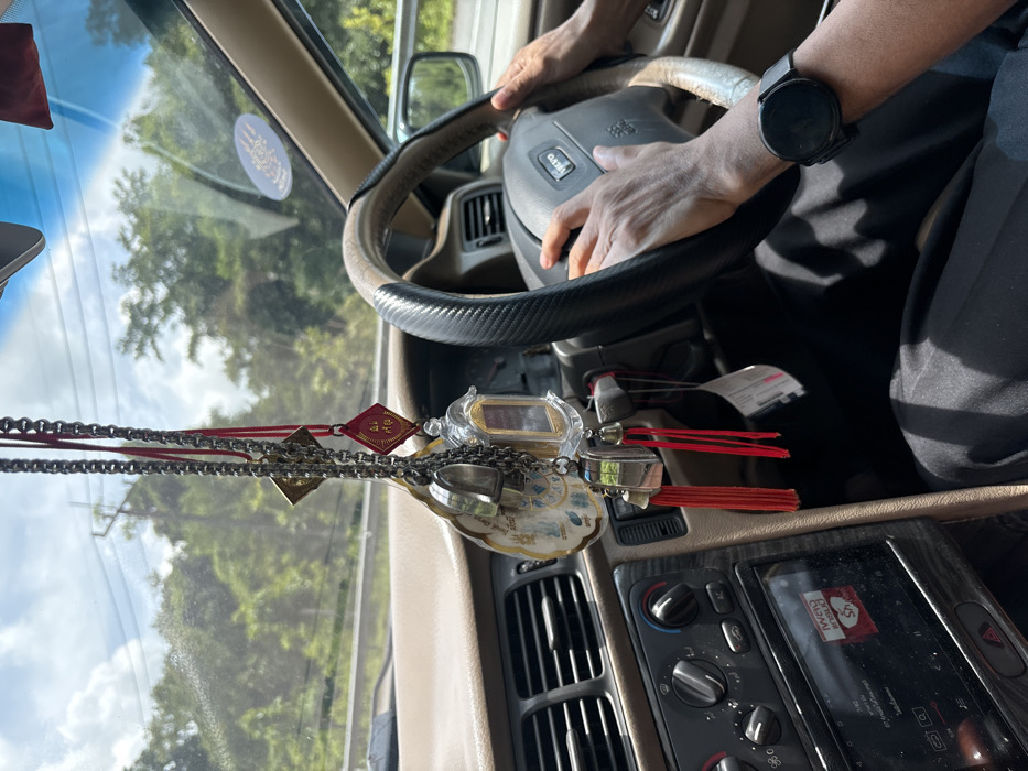 The image depicts the interior of a Volvo vehicle in motion, showing a driver's hands on the steering wheel, with a view of the dashboard and the exterior environment, located in Ban Huai To, Thailand.
**Scene:** The perspective is from the front passenger seat, looking towards the driver and the road ahead. Sunlight casts shadows within the car, indicating a bright day.
**People:** The hands and forearms of a person are visible gripping a black and tan steering wheel. The left wrist wears a dark, circular smartwatch. The person is wearing dark clothing.
**Objects (Interior):**
* **Vehicle:** The steering wheel prominently displays "VOLVO" and its logo. The dashboard is a light tan or beige color, contrasting with a darker central console.
* **Dashboard Features:** The central console includes air conditioning vents, climate controls with rotary knobs, and a modern, touchscreen infotainment system displaying Thai script and a red heart icon.
* **Hanging Ornaments:** Multiple items hang from the rearview mirror area. These include:
* Several metallic chains adorned with small silver-colored bells and charms.
* A diamond-shaped red amulet with gold Thai script.
* A small, clear, shrine-like case containing an object, suspended by a red cord.
* A larger, flat, oval-shaped amulet or charm with light blue and white imagery (possibly a Buddhist yantra) and small bells attached.
* Several red tassels dangle from these ornaments.
* **Windshield Sticker:** A circular sticker, likely an emblem or protective symbol with intricate details, is affixed to the upper right corner of the windshield.
**Environment (Exterior):**
* **Landscape:** Through the windshield, a lush, green, heavily forested, or mountainous landscape is visible under a partly cloudy blue sky. This suggests a rural or natural setting.
* **Roadside:** A white guardrail or barrier runs alongside the road on the right side of the frame, indicating a roadway.
**Actions/Interactions:** The driver's hands are actively gripping the steering wheel, suggesting the vehicle is in operation.