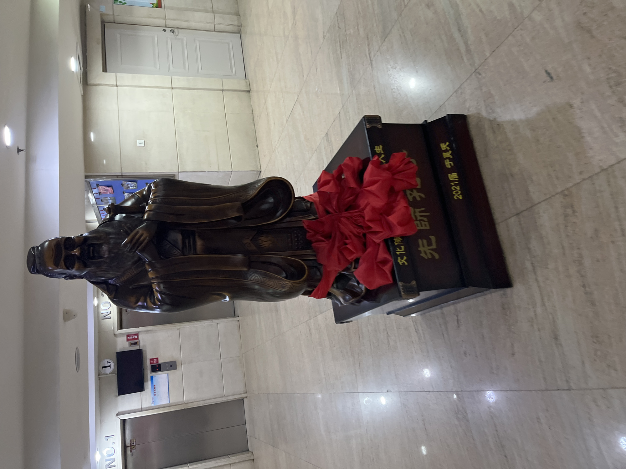 A bronze statue of Confucius stands in a building, potentially an educational or cultural institution, located in Changchun City, China. The statue is positioned on a dark brown wooden pedestal adorned with a large red bow. The pedestal bears gold lettering in Chinese characters, likely an inscription related to Confucius or the statue itself. The surrounding environment is an indoor space with tile flooring and white walls. An elevator is visible in the background, with signage indicating it is on the 1st floor. A door is also visible in the background.