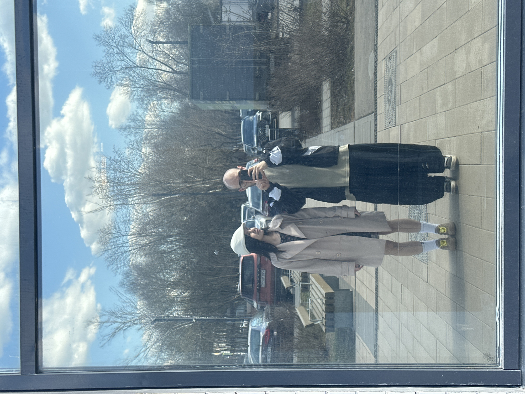 A reflection in a glass window shows two individuals standing on a paved surface in Lyubertsy, Russia. The reflected figures include a person in a long beige coat and a white hat, and another in a black jacket and dark pants, holding a phone up as if taking a picture. In the background, visible through the reflection, are bare trees, several cars, a bus stop, and a building. The sky with clouds is also reflected in the glass.