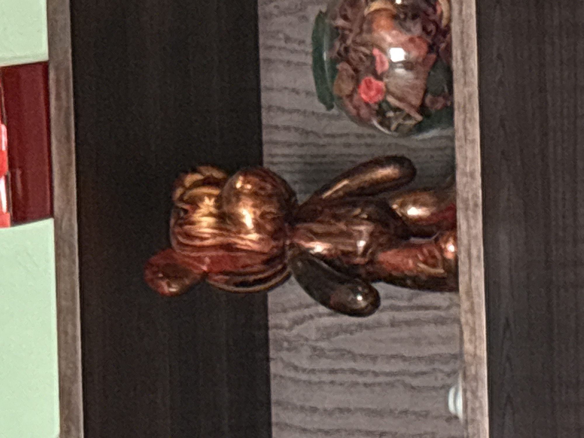 The image shows a display cabinet with a dark-colored wooden frame. Inside the cabinet, there is a metallic, copper-colored balloon animal shaped like a rabbit, lying on a shelf with a light gray woodgrain pattern. Above the rabbit, there is a clear glass bowl filled with dried flowers and leaves. The location and specific details are unknown.