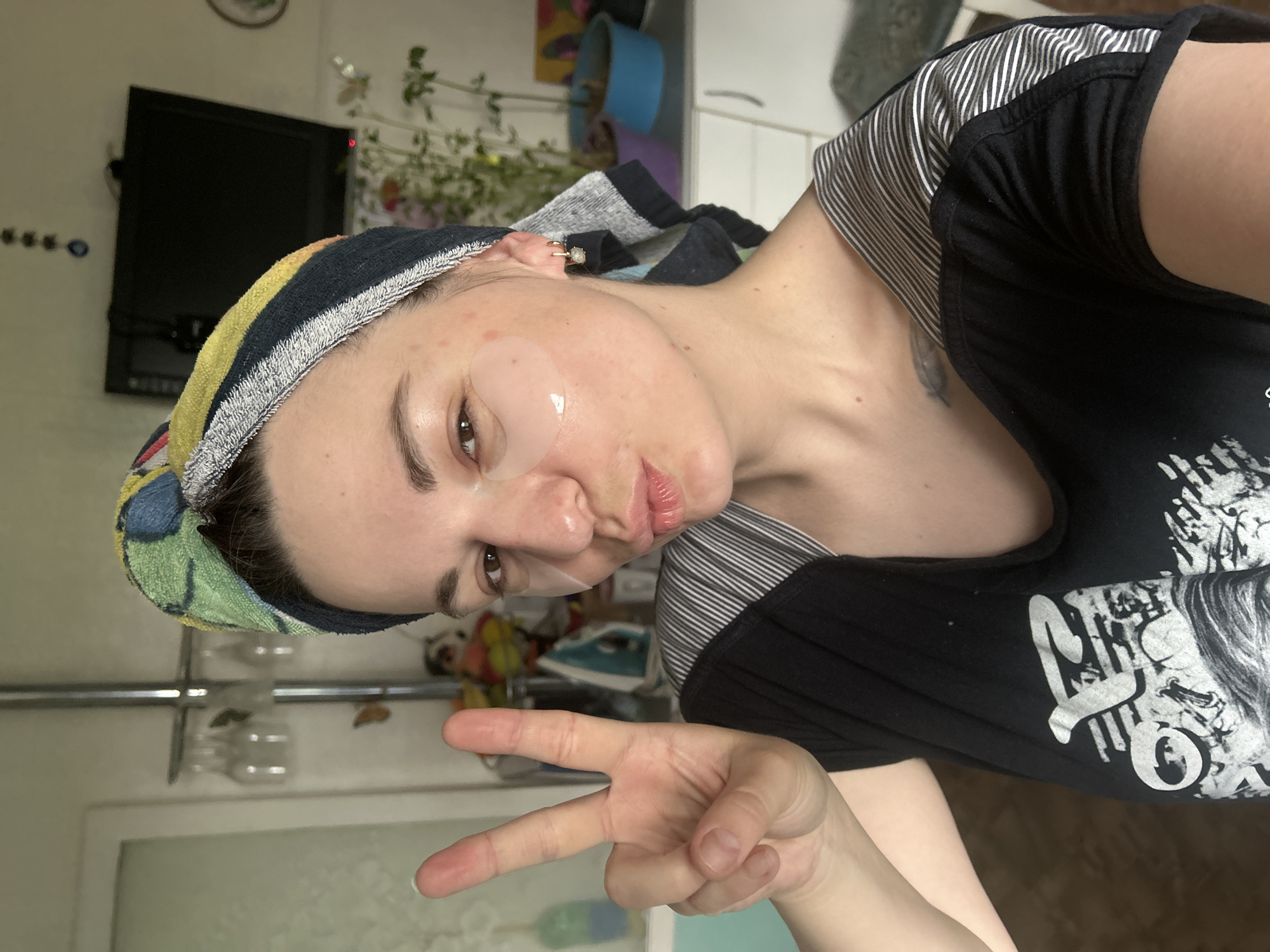 A woman in a black shirt and a colorful towel on her head is posing indoors with a peace sign. She has a translucent under-eye patch on her face. The background includes a TV, plants, a kitchen counter, and other household items. The location is Фоки, Russia.