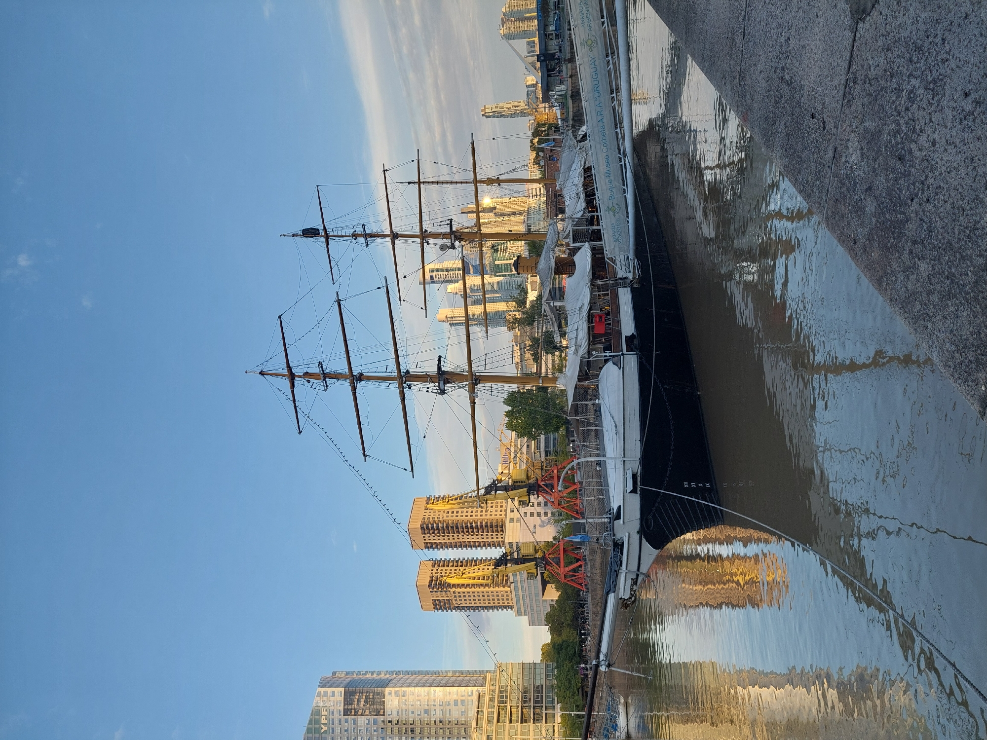 The image captures a tall ship docked at Puerto Madero, Buenos Aires, Argentina. The ship's masts stretch high against a clear blue sky with a few wispy clouds. Modern skyscrapers are visible in the background, reflecting in the water where the ship is moored. The water is murky, reflecting the buildings along the shore. The ship is docked alongside a concrete quay. No people are visible.