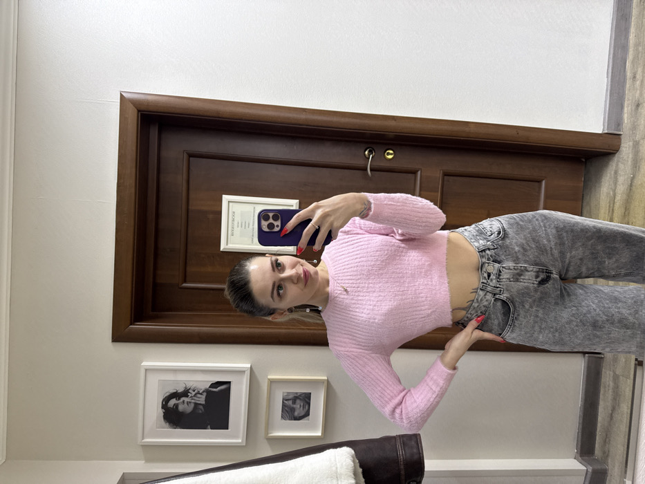 An individual is depicted taking a mirror selfie indoors in Reutov, Russia. The subject is a young adult female with dark hair pulled back, light skin, and a slight smile. She wears a pink, cropped, ribbed long-sleeve sweater, acid-wash grey high-waisted jeans, and has a visible tattoo below the hem of her sweater on her left side. Her left hand rests on her hip, displaying red nail polish and a silver-colored ring on her ring finger. Her right hand holds a dark-colored smartphone, equipped with a purple case, pointed towards the mirror. She also wears earrings and a delicate necklace.
The background consists of a light-colored, possibly cream, wall. To the left, a dark brown jacket with a white/cream fur-lined collar or interior hangs on a rack. Above it, two framed black and white photographs are mounted vertically: the upper one depicts a woman with dark hair smoking, and the lower shows a woman's face. Directly behind the subject, a dark brown wooden door with a brass-colored handle and lockplate is visible. A white-framed document or certificate, featuring Cyrillic text (e.g., "АТТЕСТАТ" at the top), is affixed to the door. The floor appears to be a light-colored wood or laminate. The scene is illuminated by ambient indoor lighting.