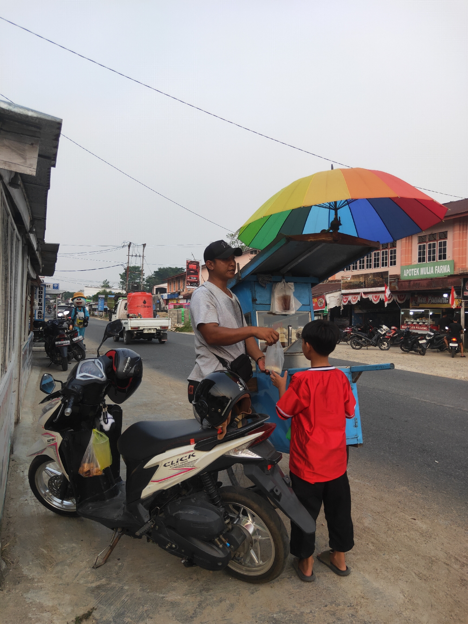 A man wearing a grey t-shirt and a black baseball cap is selling food from a cart under a colorful umbrella on a street in Sintang, Indonesia. A young boy in a red t-shirt and black pants stands facing the man, holding a plastic bag. The cart is blue with white trim and has a metal railing. Two motorcycles are parked in the foreground, one white and black, the other black. A yellow plastic bag containing food is hanging from the handlebars of the white and black motorcycle. In the background, there are buildings, including a pharmacy with a pink facade and green signage that reads "APOTEK MULIA FARMA." The sky is overcast. The scene appears to be on a commercial street with shops and businesses lining the road.
