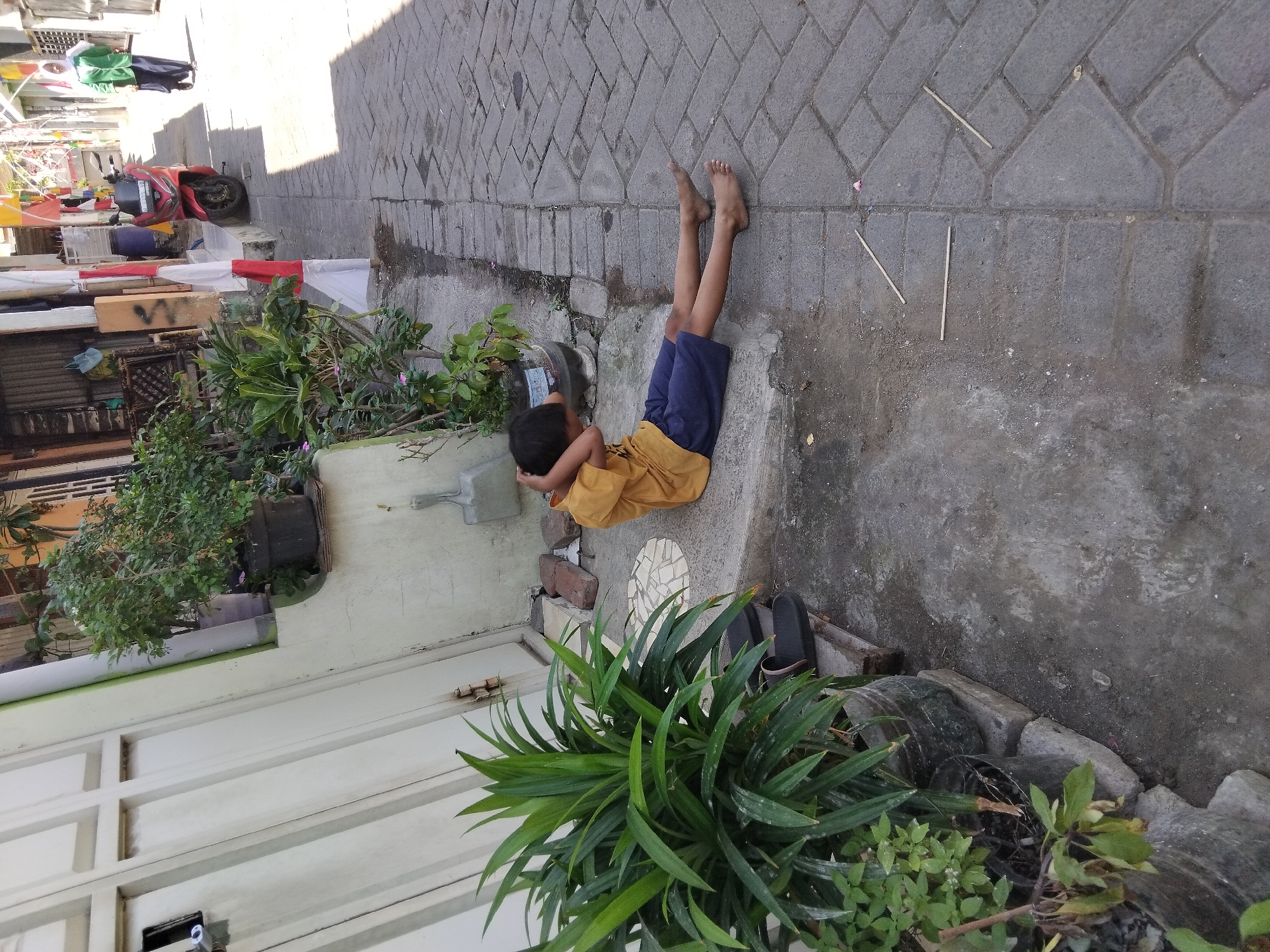 A young boy in a yellow t-shirt and blue shorts is sitting on the ground with his legs extended on a paved walkway. He is resting his head on his hands, with his elbows bent and his hands behind his neck. To his left is a large potted plant with long, green leaves. Behind the plant is a closed, light-colored door or gate. The walkway is made of interlocking gray pavers. Further down the walkway, a red motorcycle is parked. In the background, a person wearing a green jacket and headscarf is walking away from the camera. The scene appears to be in a residential area with buildings lining the walkway. The lighting suggests it is daytime.