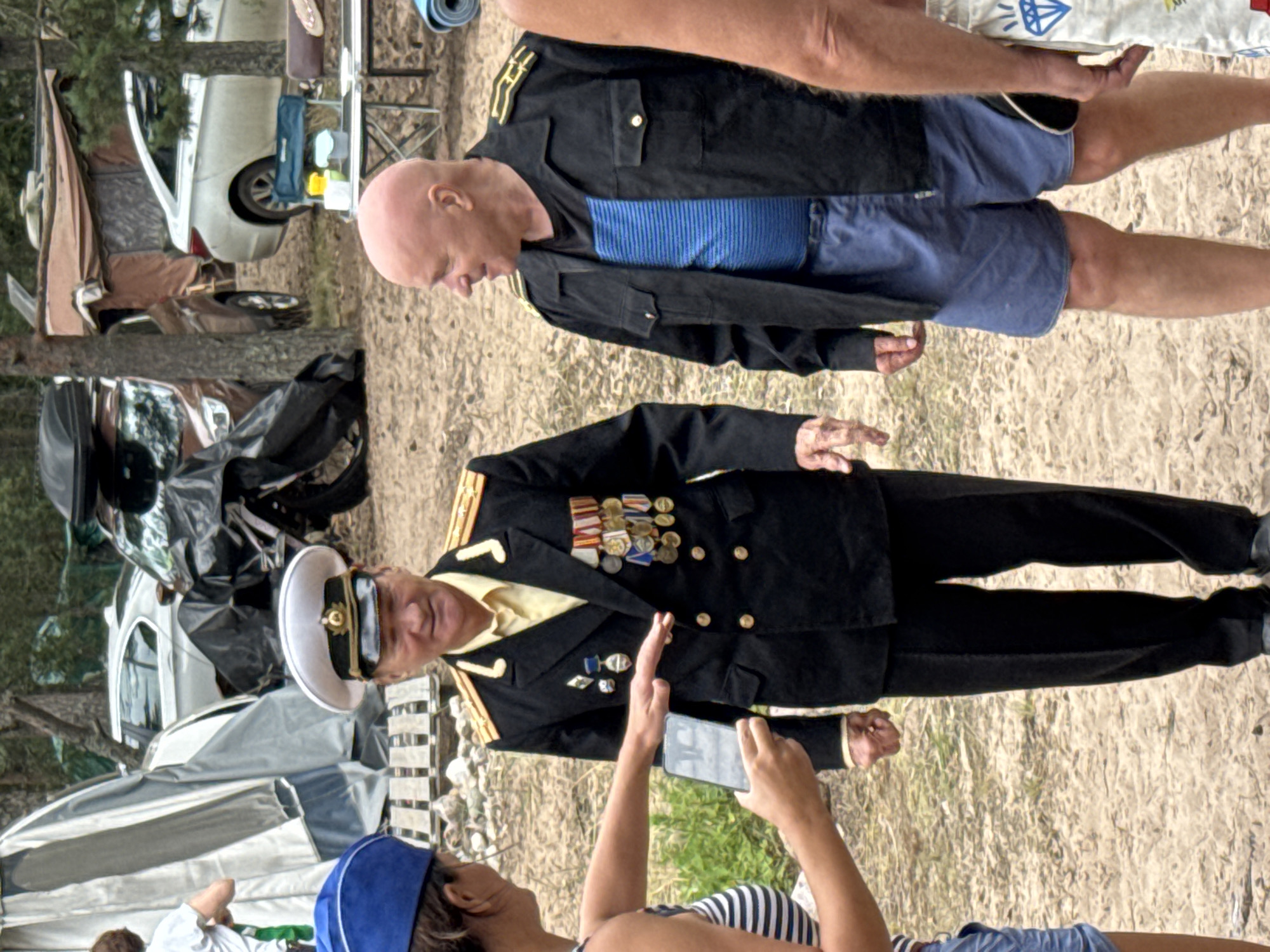 In Zelenaya Roshcha, Russia, a medium shot captures a sandy outdoor environment with sparse trees and some scattered tents and vehicles. Three individuals are the primary subjects. On the left, a woman wearing a blue hat and a striped top is seen from the chest up, partially interacting with a man to her right. Her hand is extended, holding a smartphone, as if taking a photo or video.
The central figure is a man in a formal black military-style uniform with gold epaulets, a white cap, and numerous medals displayed on his chest. He is facing forward with a slight smile. To the right of the central figure is another man, bald and wearing a black jacket over a blue striped shirt and blue shorts. He is also facing forward, looking slightly to his left.
In the background, a white car is visible, along with parts of tents and what appears to be a motorcycle with a black cover. The ground is covered in sand and dirt. The lighting suggests it is daytime, likely a warm season given the attire of the individuals.