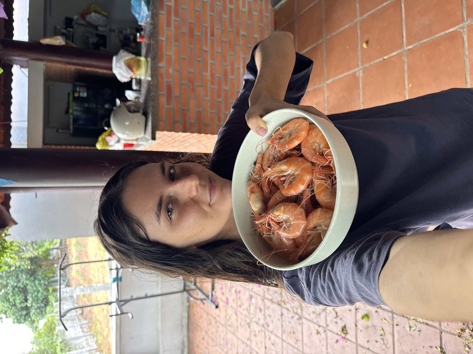 A woman is holding a bowl of cooked shrimp in what appears to be an outdoor covered patio area in Mui Ne, Vietnam. The patio has terracotta tiled flooring and brick walls. In the background, there is a refrigerator, shelves stocked with items, and a white helmet on a counter. Lush green trees and a chain-link fence are visible beyond the patio. The woman is looking directly at the camera with a slight smile.