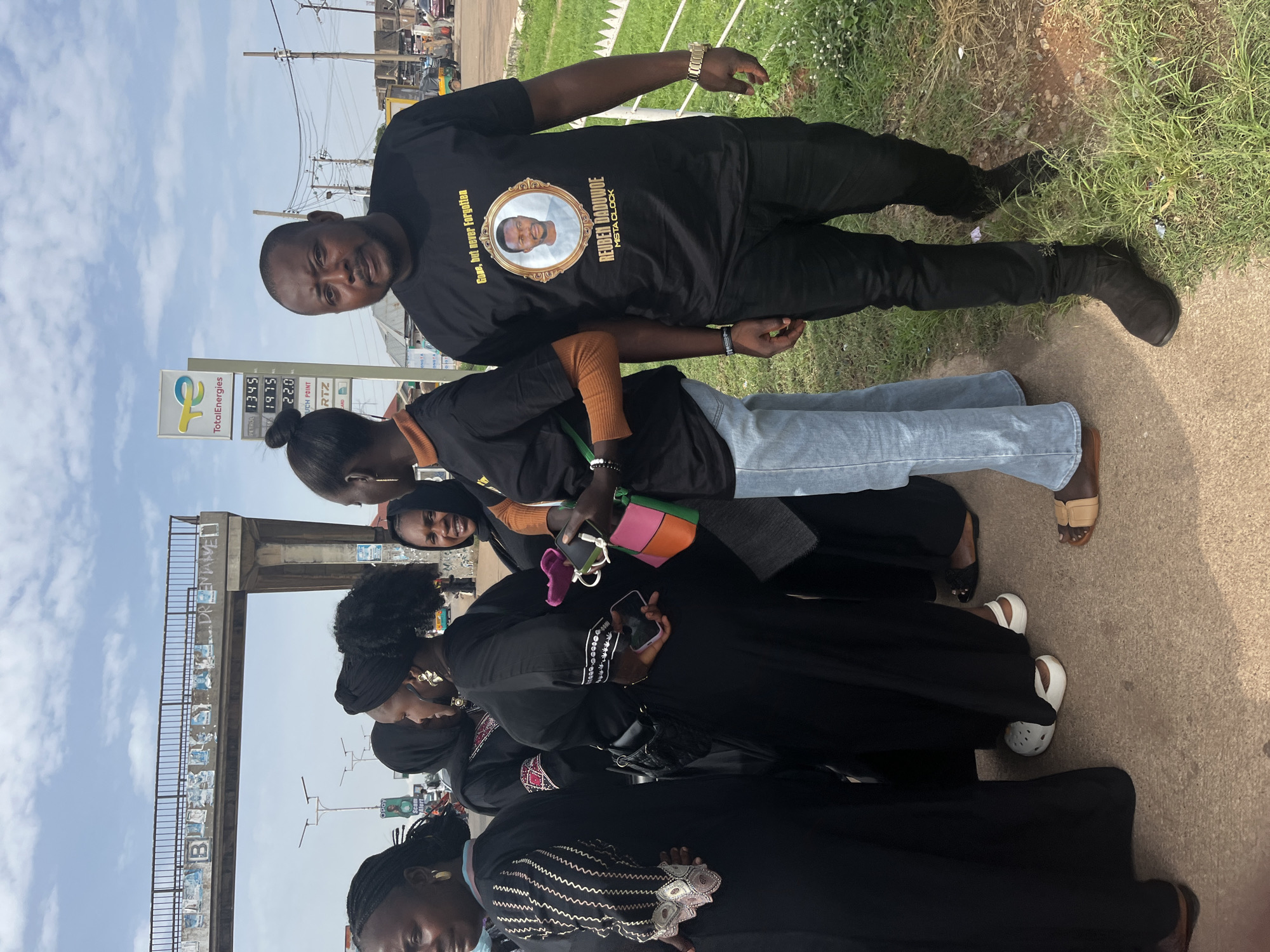 The image captures a group of individuals standing near a TotalEnergies gas station in Gura Top, Nigeria. In the foreground, a man wears a black t-shirt with a memorial image of a man named Reuban Daduboe, also known as Msta Clock, with the text "Gone, but never forgotten." He is accompanied by several women dressed in black attire, some with headscarves. The background features a pedestrian bridge with graffiti, a gas price display, and other buildings and vehicles suggesting an urban environment. The overall atmosphere appears somber, likely related to the memorial on the man's t-shirt.