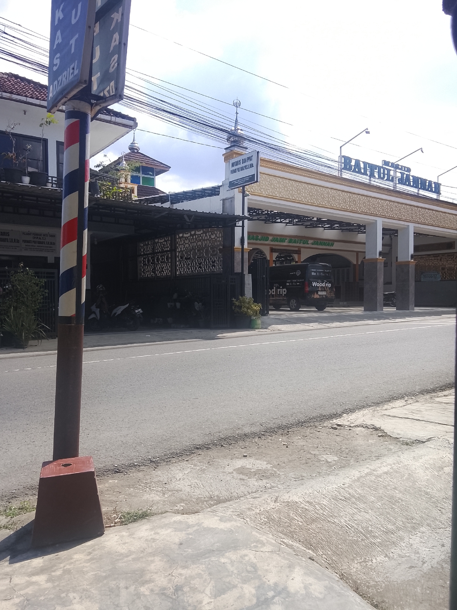 The image depicts the facade of a building identified as "MASJID JAMI BAITUL JANNAH" in Karangklesem, Indonesia. The building features an arched entryway with decorative gold accents and pillars. A black van with the text "Woodrip" is parked in front of the building. To the left, a barber pole and a sign with text in Indonesian, including "KUST ADZRIEL," are visible. The scene is set on a street with a sidewalk and overhead power lines. The sky is clear and blue, suggesting it is daytime.