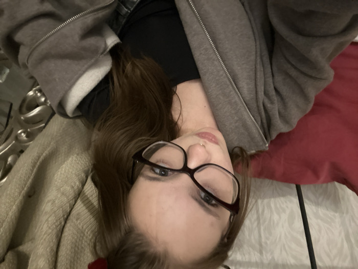 A person with brown hair and glasses is lying on their back. They are wearing a black top and a gray zip-up hoodie. Part of a decorative silver bedframe and a tan blanket are on the left. A red pillow and a white sheet with a dark bar across it are on the right. The person has fair skin and a piercing in their nose.