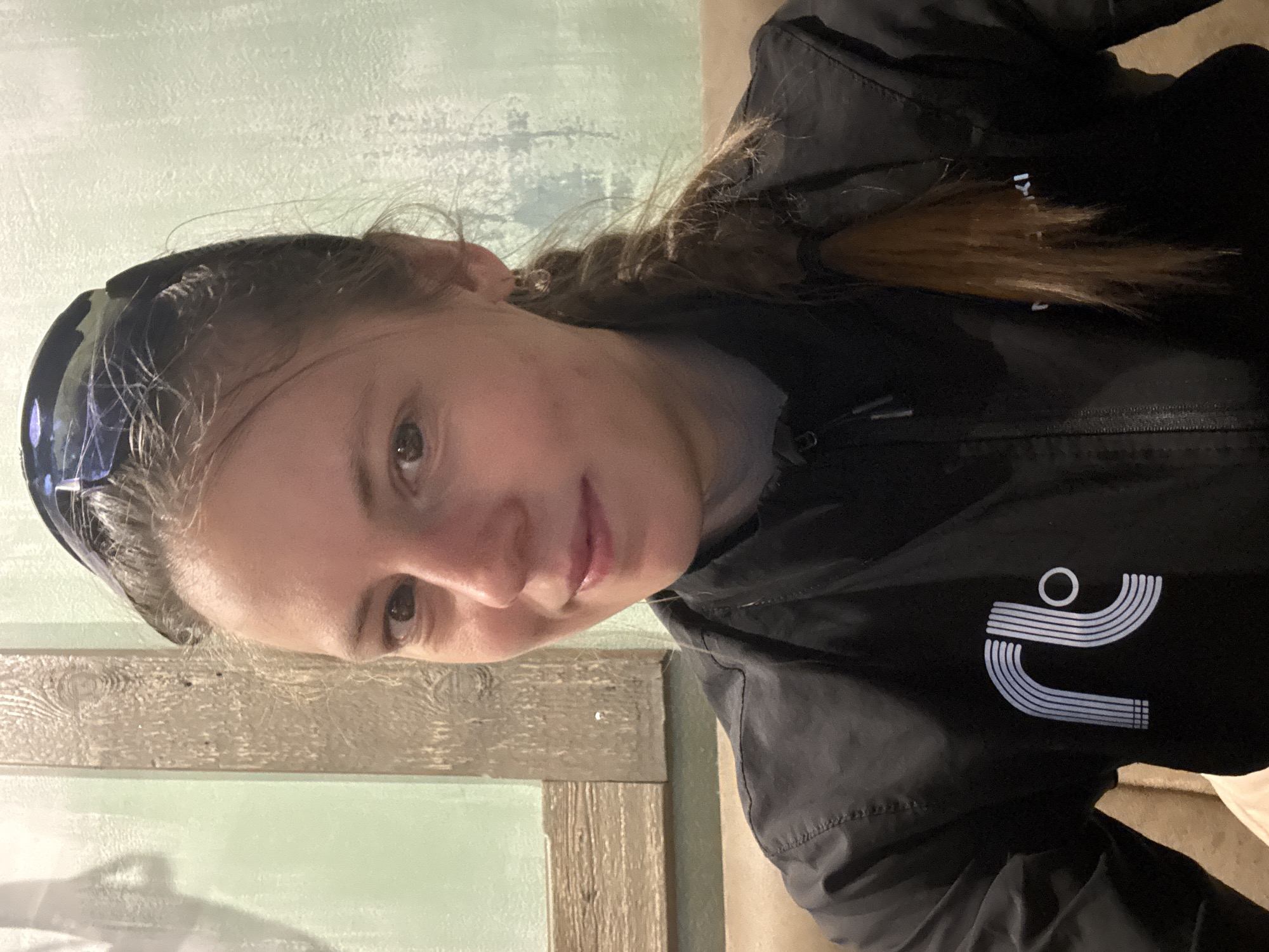 A young woman is photographed indoors, likely in Kazan, Russia. She is wearing a black jacket with a white logo on the right breast. Her hair is pulled back in a braid and she has a headband on her head. The background consists of a light green wall and a wooden frame.