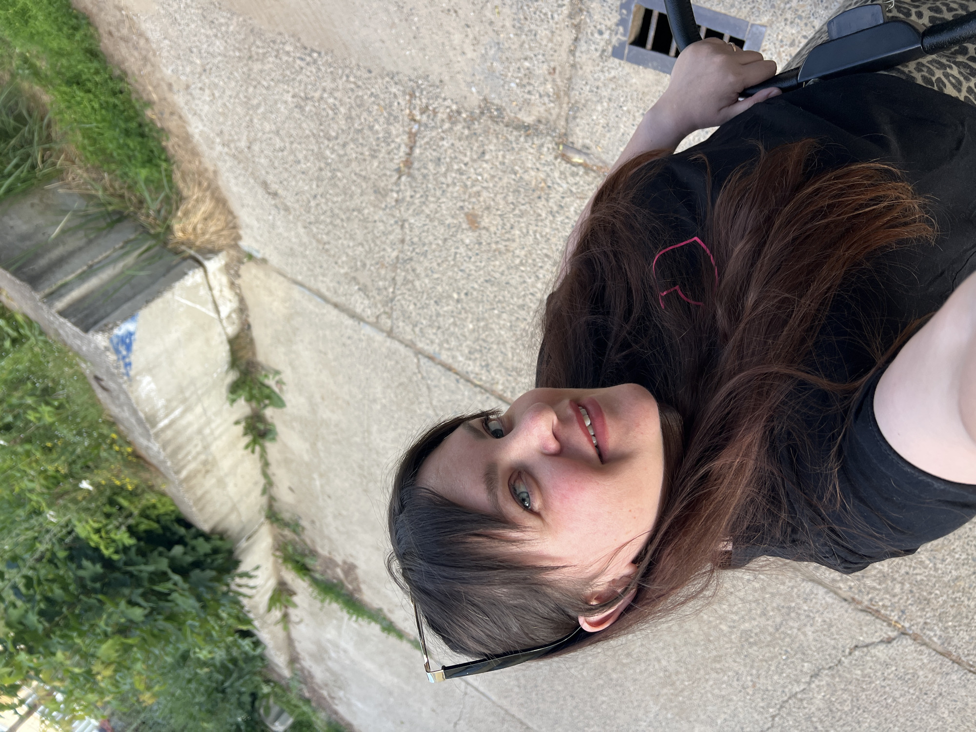 A young woman with long, dark brown hair and blue eyes smiles at the camera. She is wearing a black t-shirt with a pink heart design and appears to be outdoors, seated on or near a stroller. Her eyeglasses are resting on her t-shirt. The background consists of a concrete surface, grass, and a drainage grate. The lighting suggests it is daytime.