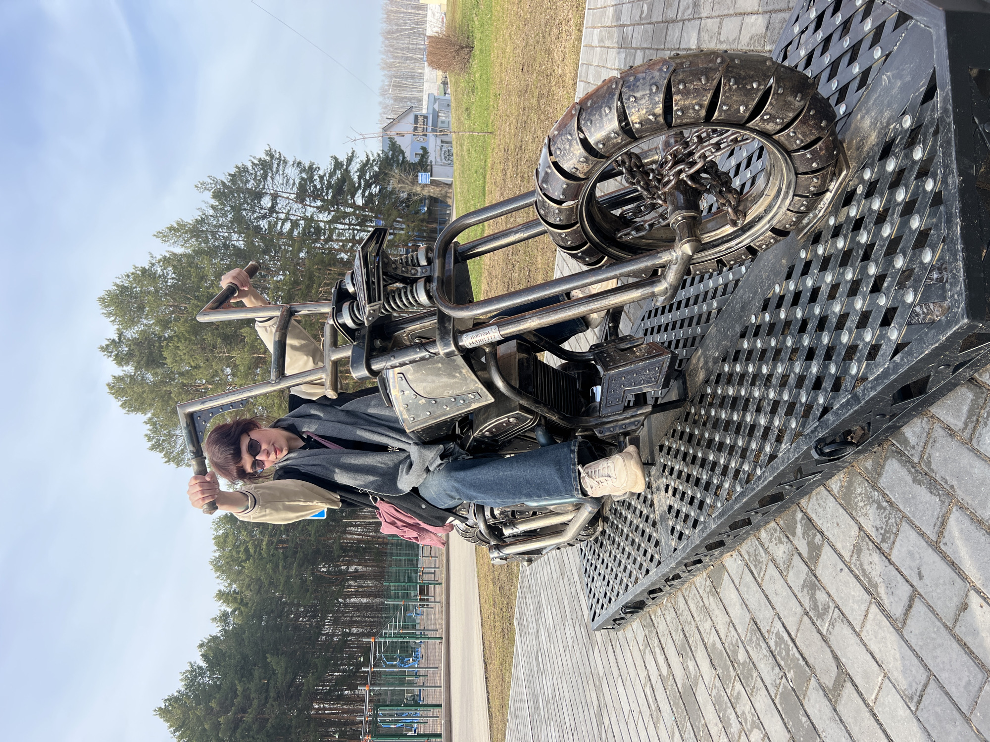 A person is sitting on a metal motorcycle sculpture located in the city of Chaikovsky, Russia. The individual is wearing sunglasses, jeans, and a jacket. The motorcycle is made of metal and has a distinctive, chunky wheel. The sculpture is positioned on a raised metal platform with a perforated pattern. The background includes a park area with trees, green grass, and a fence. Buildings and a cloudy sky are visible in the distance. The foreground consists of paving stones.