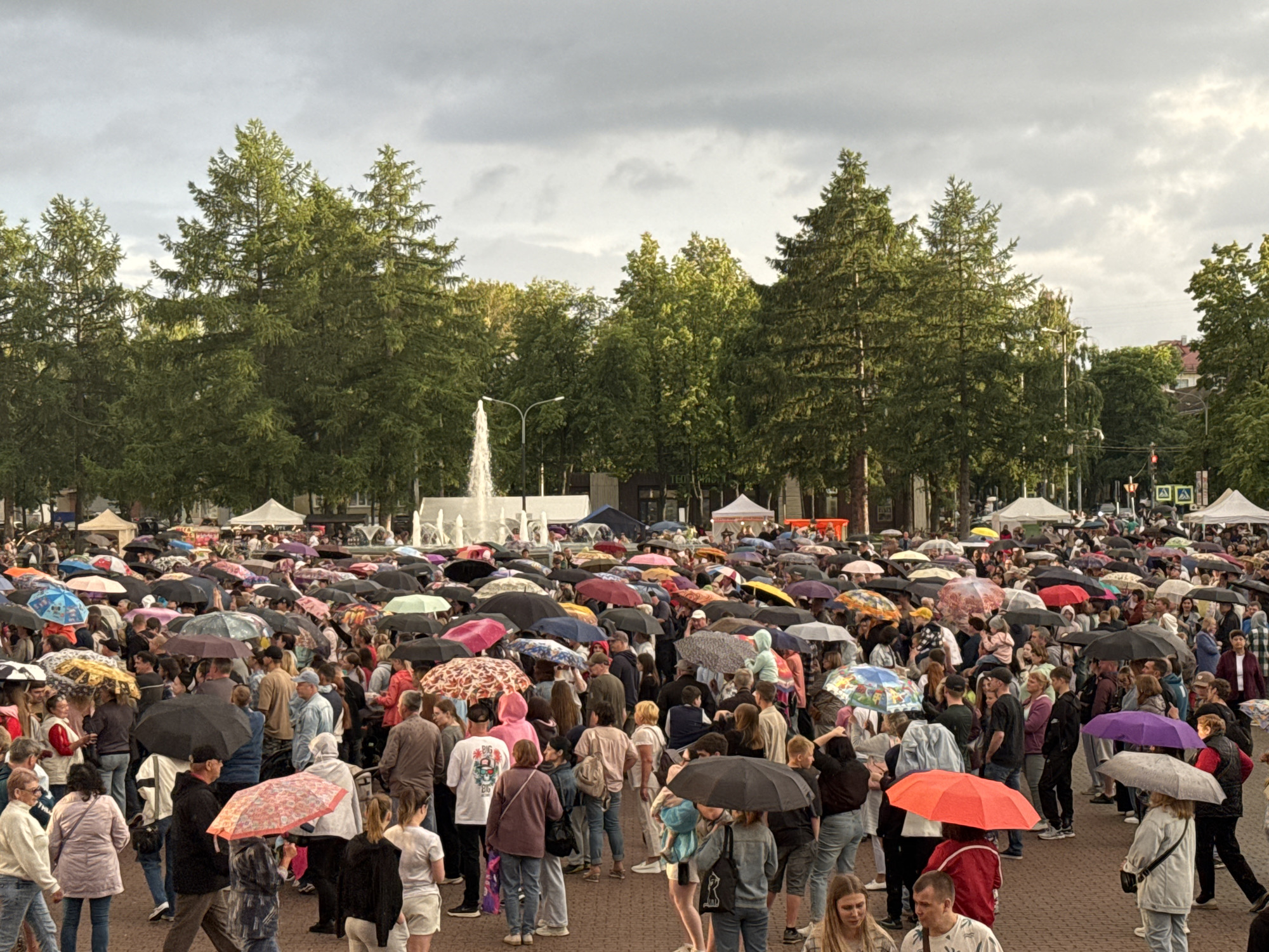 The image depicts a crowded outdoor public space in Chaikovsky, Russia, under a cloudy sky, with many people holding umbrellas, suggesting it is raining or has recently rained. The crowd is dense, with individuals of various ages and genders, dressed in casual attire. Many are looking in the same general direction, possibly towards a focal point not entirely visible, or simply as part of the gathering. The environment features numerous trees, a fountain in the background, and some temporary structures like tents or canopies. The ground appears to be paved. The overall atmosphere is that of a public event or gathering taking place despite the weather.