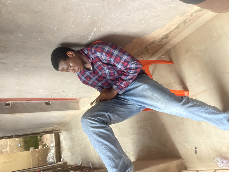 A young, dark-skinned man sits on an orange chair, leaning forward slightly. He wears a red, blue, and white plaid shirt with blue jeans. He has short, dark hair. The background includes a worn, painted wall and ceiling. A window shows an exterior view of green foliage and scattered refuse. The perspective is unusual, as if the image was rotated.