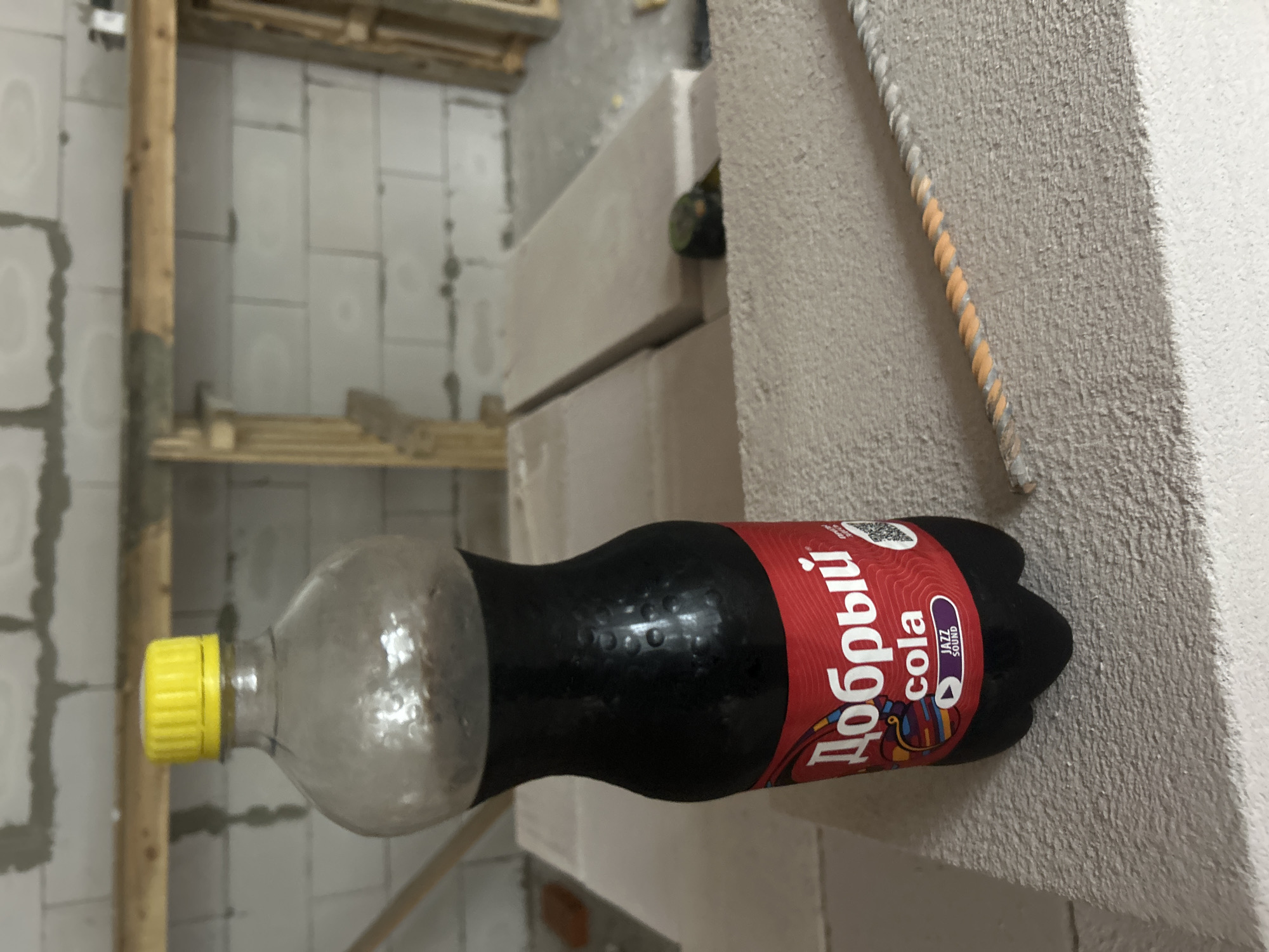 A plastic bottle of "Добрый" cola with a yellow cap sits on a rough, light-colored surface. The bottle has a red label with white text that reads "Добрый cola" and a smaller purple label that says "JAZZ SOUND". The bottle is partially filled with a dark liquid. In the background, a wall constructed of light-colored, rectangular blocks is visible, with wooden beams and framing elements. A section of rebar with orange and gray wrapping lies diagonally across the surface in the foreground. The environment appears to be an unfinished construction site. There are no people present in the image.