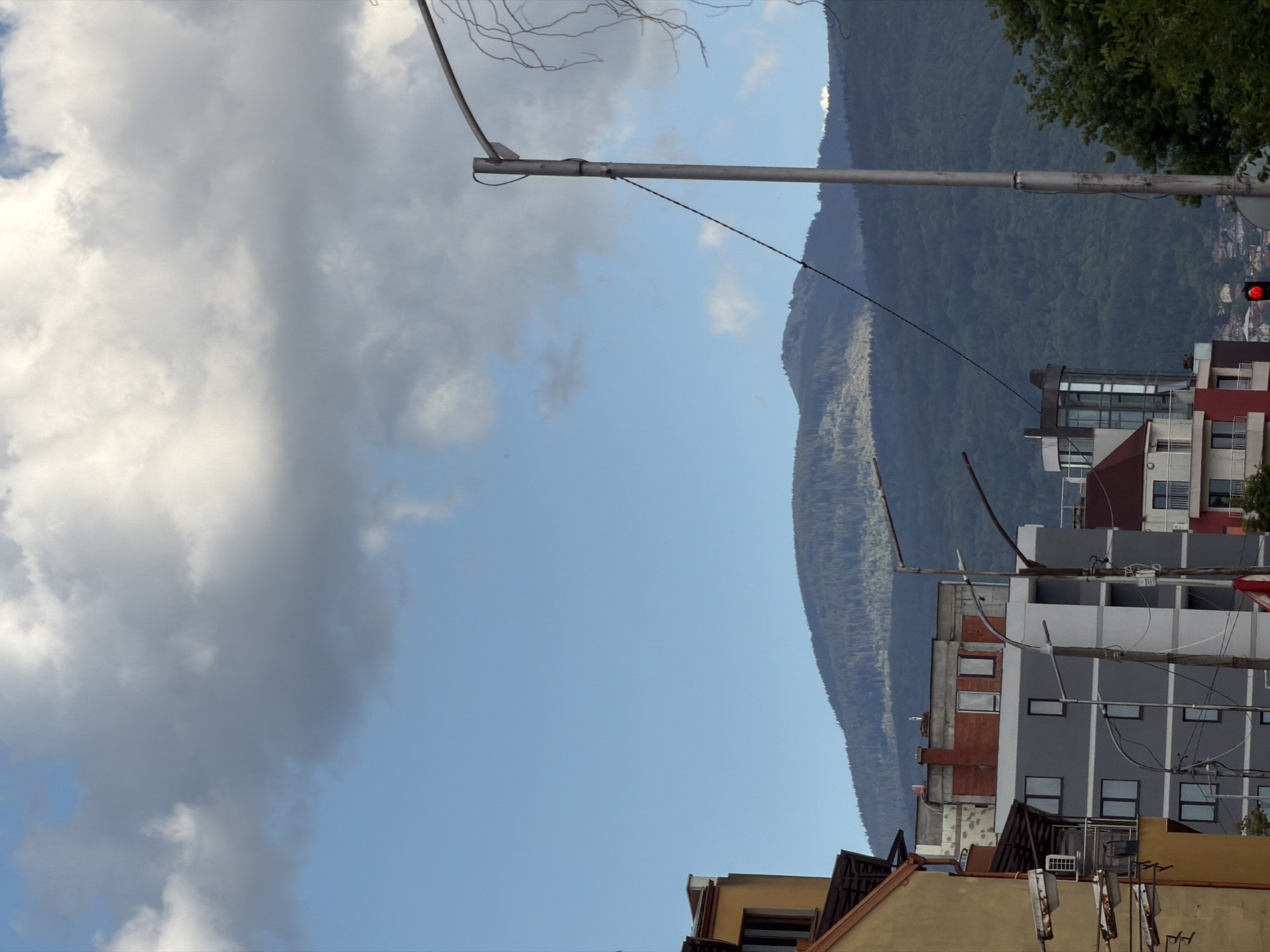 The image shows a view in Sofia, Bulgaria, featuring a mountain range in the background. A gray metal light post extends across the top of the frame. Below the light post, there are various buildings of different heights and colors. Some are white and gray, while others have red or brown elements. A red traffic light is visible on one of the buildings. The sky transitions from light blue to gray, with large clouds on the left side of the image. The overall environment includes urban elements and natural scenery.