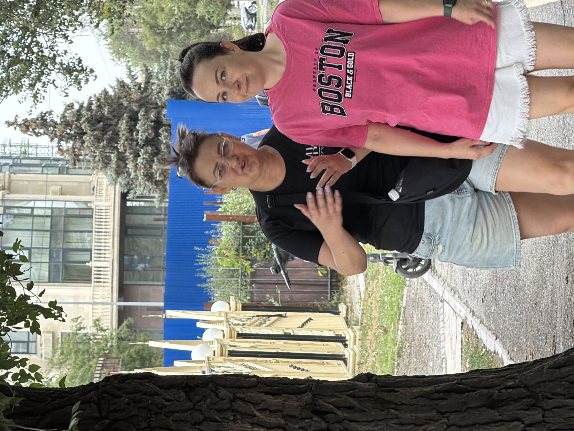 Two women walk along a paved path next to a tree. The woman on the left wears glasses, a black t-shirt, and denim shorts. She gestures with her hands as she speaks. The woman on the right wears a bright pink t-shirt with "BOSTON BLACK & GOLD" printed on it, and white fringed shorts. She looks at the camera. In the background, a building with a balcony and a blue fence are visible. A scooter is parked on the side of the path.