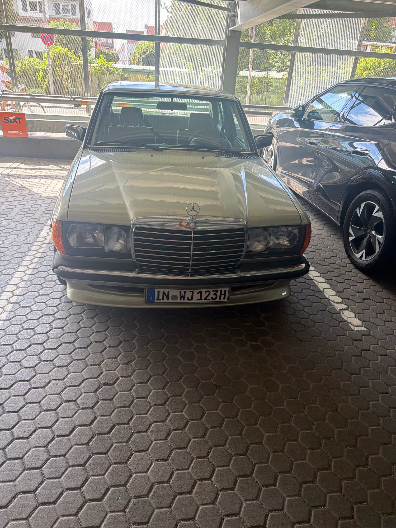 The image depicts a light-colored Mercedes-Benz W123 sedan parked in a covered parking area with hexagonal patterned paving stones. The car's front grille, headlights, and license plate, which reads "IN WJ 123H," are clearly visible. A darker SUV is parked next to the Mercedes. In the background, through a metal mesh fence, a street scene with buildings and trees is visible. A red and white "SIXT PARKING" sign is mounted on a railing to the left of the Mercedes. The scene appears to be in Ingolstadt, Germany, suggested by the license plate.