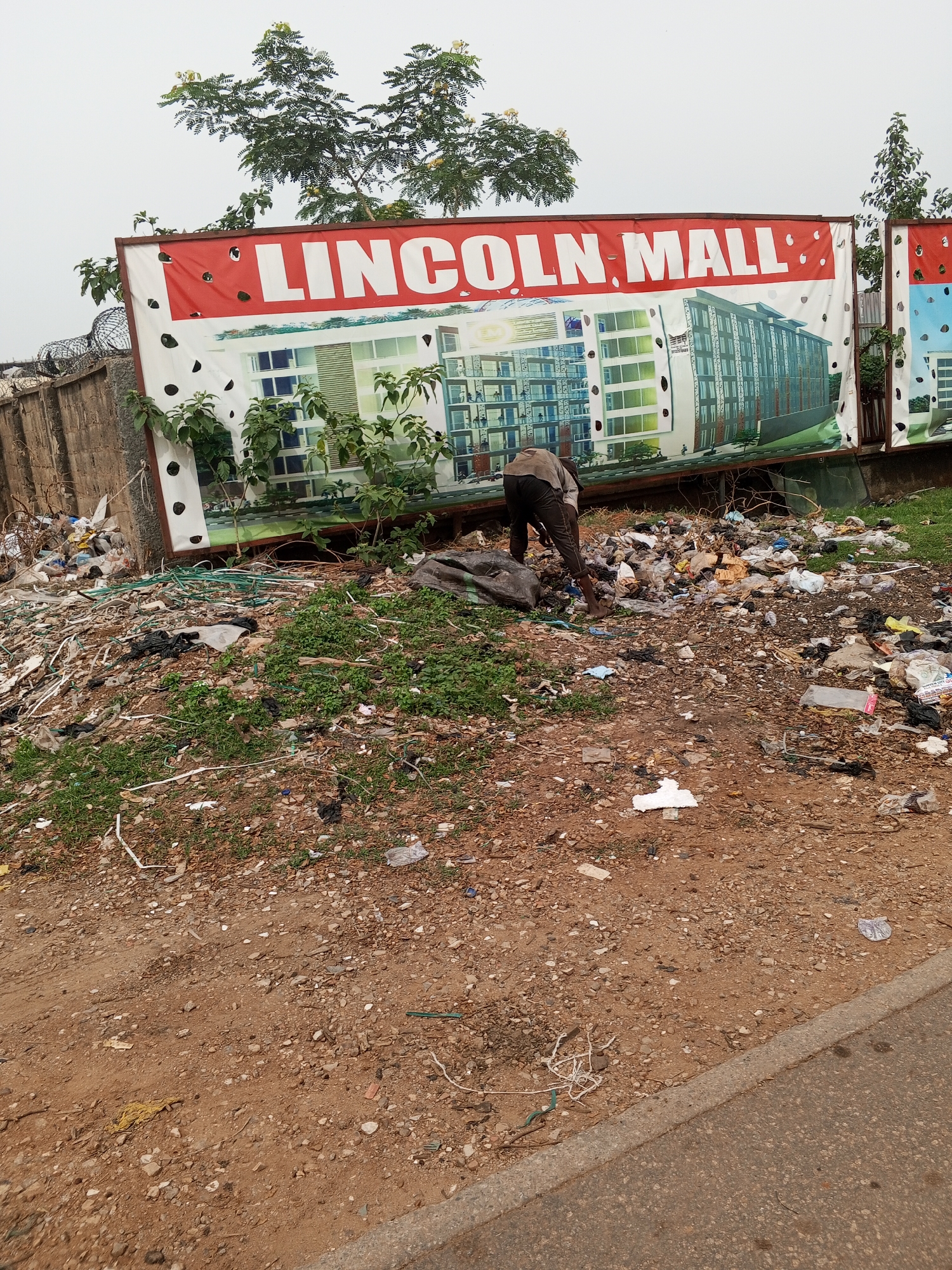 A man scavenges through a large pile of garbage in front of a billboard advertising "Lincoln Mall." The billboard features images of modern-looking buildings. The area is littered with trash, and there is a dirt ground leading up to the garbage pile. The scene appears to be in disrepair, with trash scattered around the environment. A concrete wall topped with barbed wire is visible in the background. The location is inferred to be in Abuja, Nigeria, based on the name "Lincoln Mall," which, while not necessarily exclusive to the city, is a common type of business name found in urban areas.