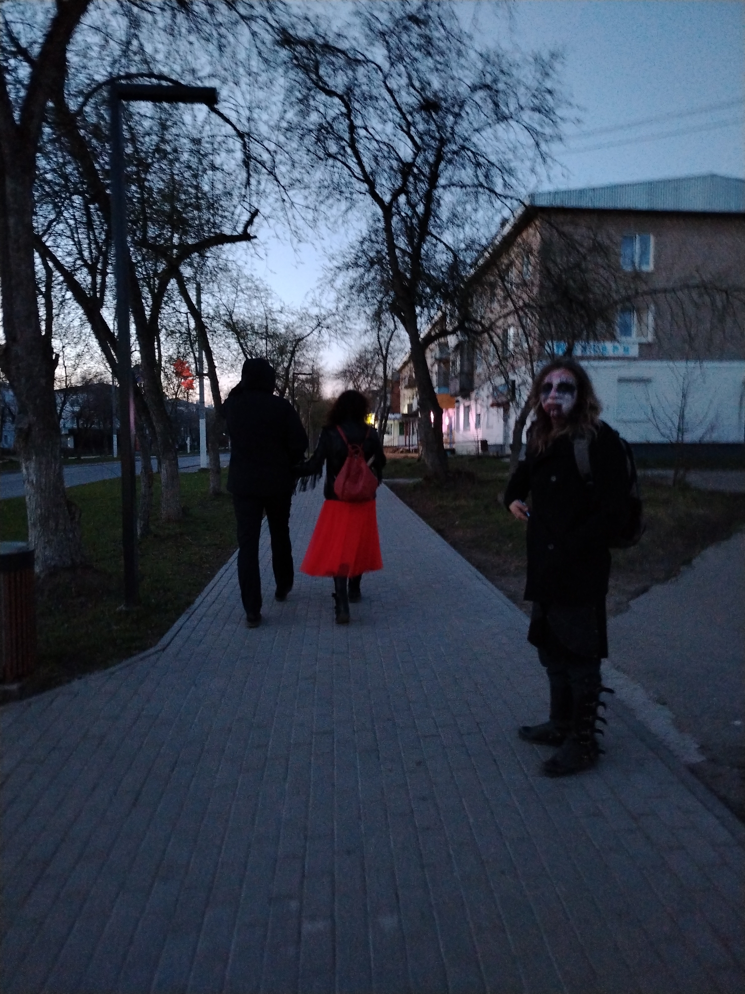 The image captures an outdoor scene in what appears to be Chaikovsky, Russia, possibly taken at dusk. In the foreground, a person with zombie-like makeup is standing on a brick-paved sidewalk. They are wearing a dark coat, carrying a backpack, and their boots have distinctive, possibly gothic, design features. To the left of this individual, a man and woman are walking away from the camera, hand in hand. The man is dressed in black while the woman is wearing a bright red skirt and a red backpack. The background consists of trees, street lights, and a multistory building with visible windows and a sign with lettering in a non-English script. The environment suggests a residential or suburban setting. The lighting is subdued, indicative of the time of day.