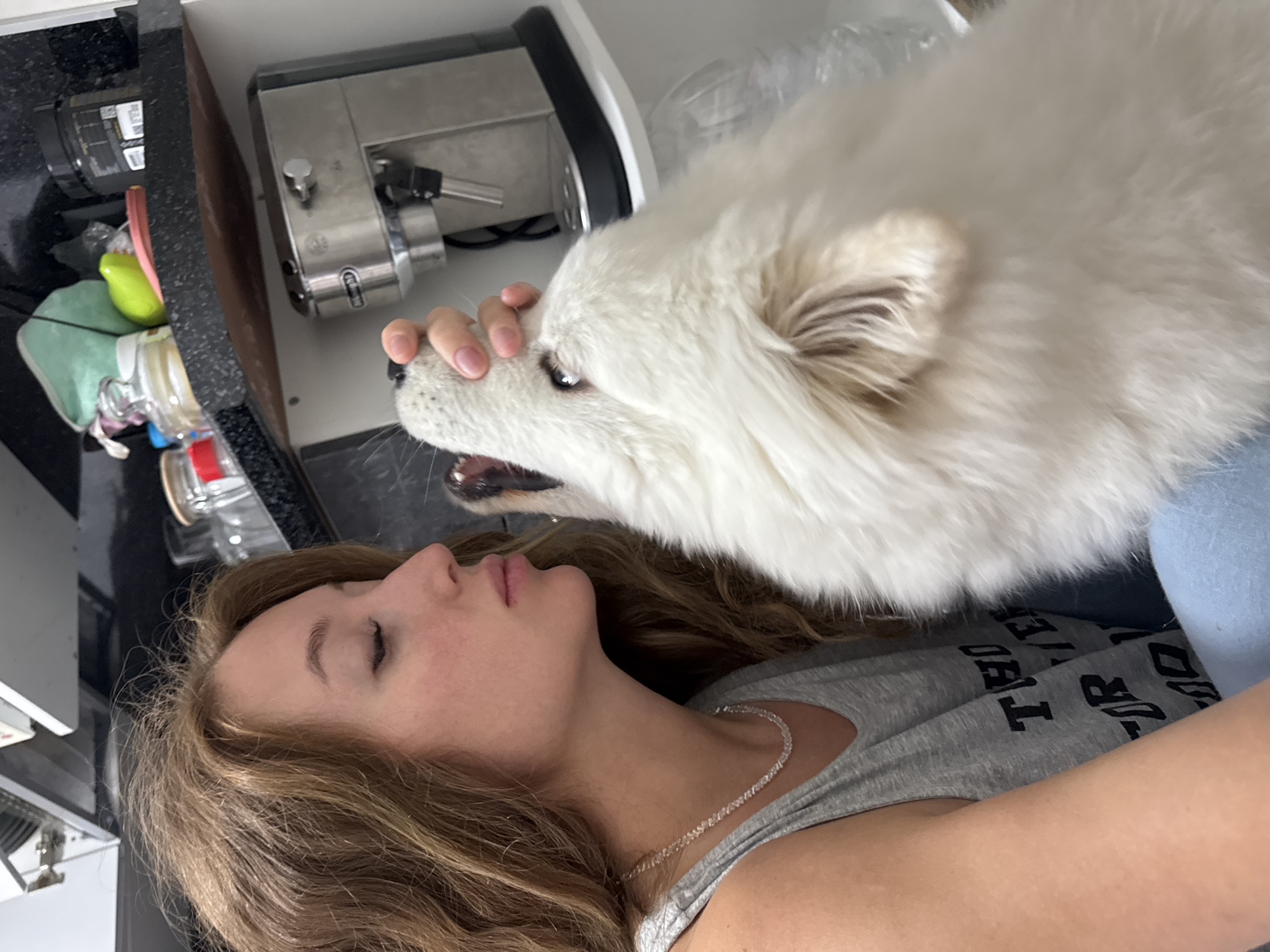A young woman lies supine, indoors. A large, white Samoyed dog stands over her, with the woman's right hand resting on the dog's snout. The woman wears a light gray t-shirt and a silver necklace. A stainless steel espresso machine, various containers, and kitchen items are on the counter behind the woman and dog. The location is unknown but not necessarily located in Chaikovsky, Russia.