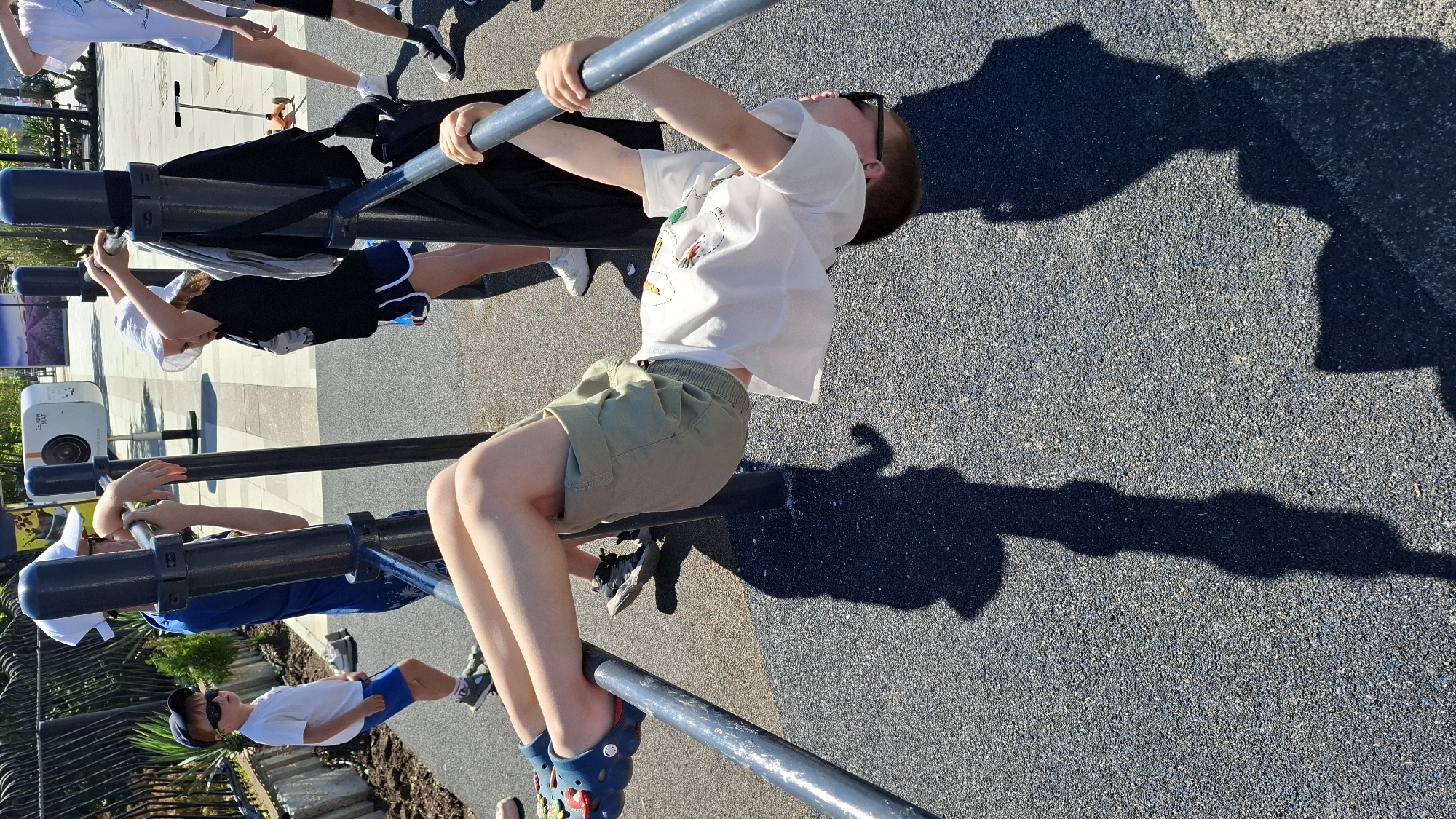 The image captures an outdoor scene with several children engaged in activity around metal exercise bars, likely in a park or playground. The lighting suggests a sunny day, with long, distinct shadows cast on the dark gray, textured paved ground.
In the foreground, a boy wearing a white t-shirt, khaki shorts, sunglasses, and blue Crocs is hanging from a horizontal metal bar, looking upwards. His body is slightly arched, indicating he is actively holding onto the bar. Behind him, another child, wearing a black t-shirt, dark shorts, and a white hat, is also hanging from a lower metal bar. Other children are partially visible in the background, some standing near the structures.
The metal bars are dark gray or black, part of a multi-station outdoor fitness or play equipment. A black bag or jacket is draped over one of the bars near the second hanging child. To the left, a white projector-like device labeled "CEDAR GEAK" and "HD PROJECTOR" is visible on a stand, possibly indicating an outdoor event or exhibit. The overall atmosphere appears casual and active, with the children enjoying the outdoor equipment.