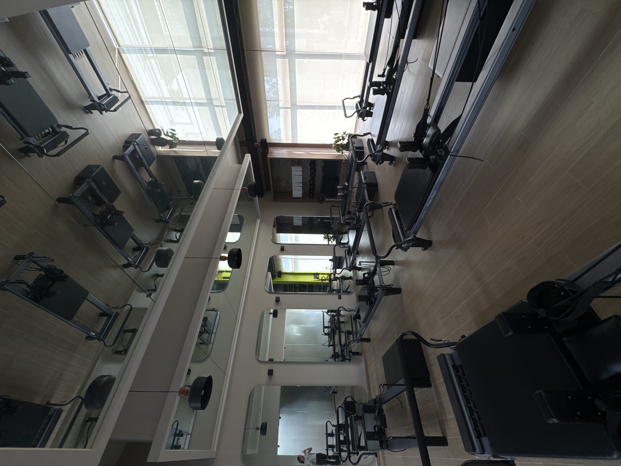 A fitness studio in San Bernardino Tlaxcalancingo, Mexico, features exercise equipment such as pilates reformers in a row. The studio has mirrors on the walls and a skylight ceiling. The floor appears to be made of wood. The studio is empty of people except for a person who is visible in the reflection of one of the mirrors.