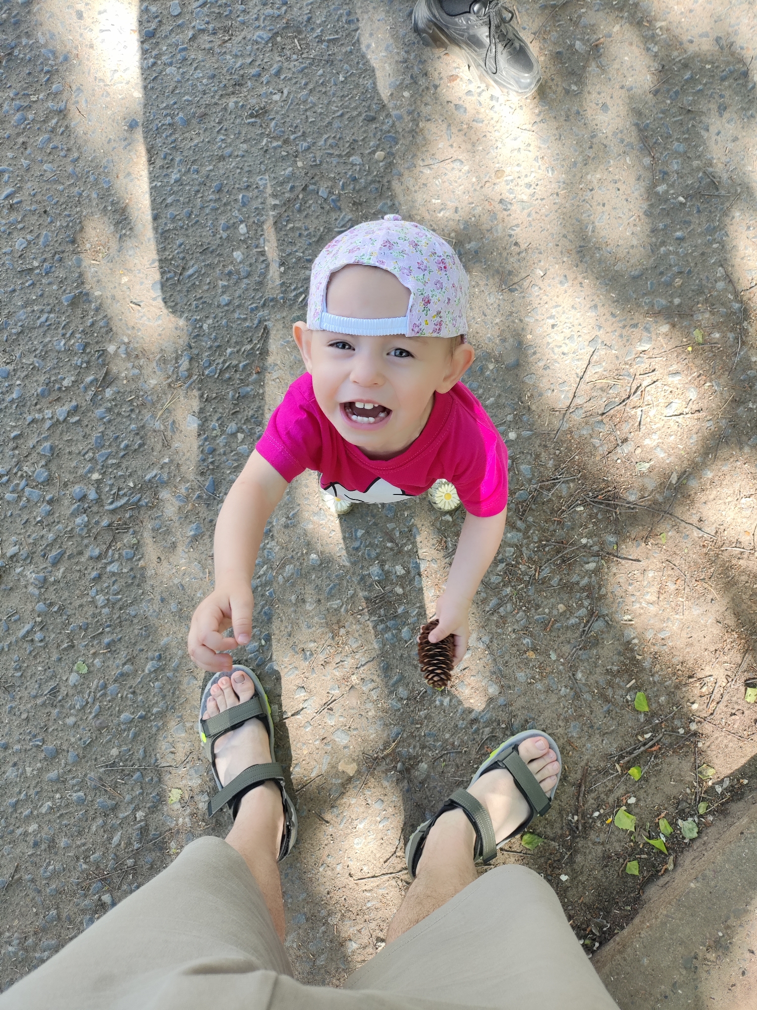 A young child wearing a floral patterned cap and a pink shirt is holding a pine cone and looking up at the camera. The child is standing on a gravel ground surface. An adult's legs and feet are visible at the bottom of the frame. The adult is wearing sandals and khaki shorts. Another adult's shoe is visible in the upper right corner of the frame. Shadowy light patterns are cast on the ground. Fallen leaves and twigs are scattered on the ground. The location is possibly Chaikovsky, Russia.