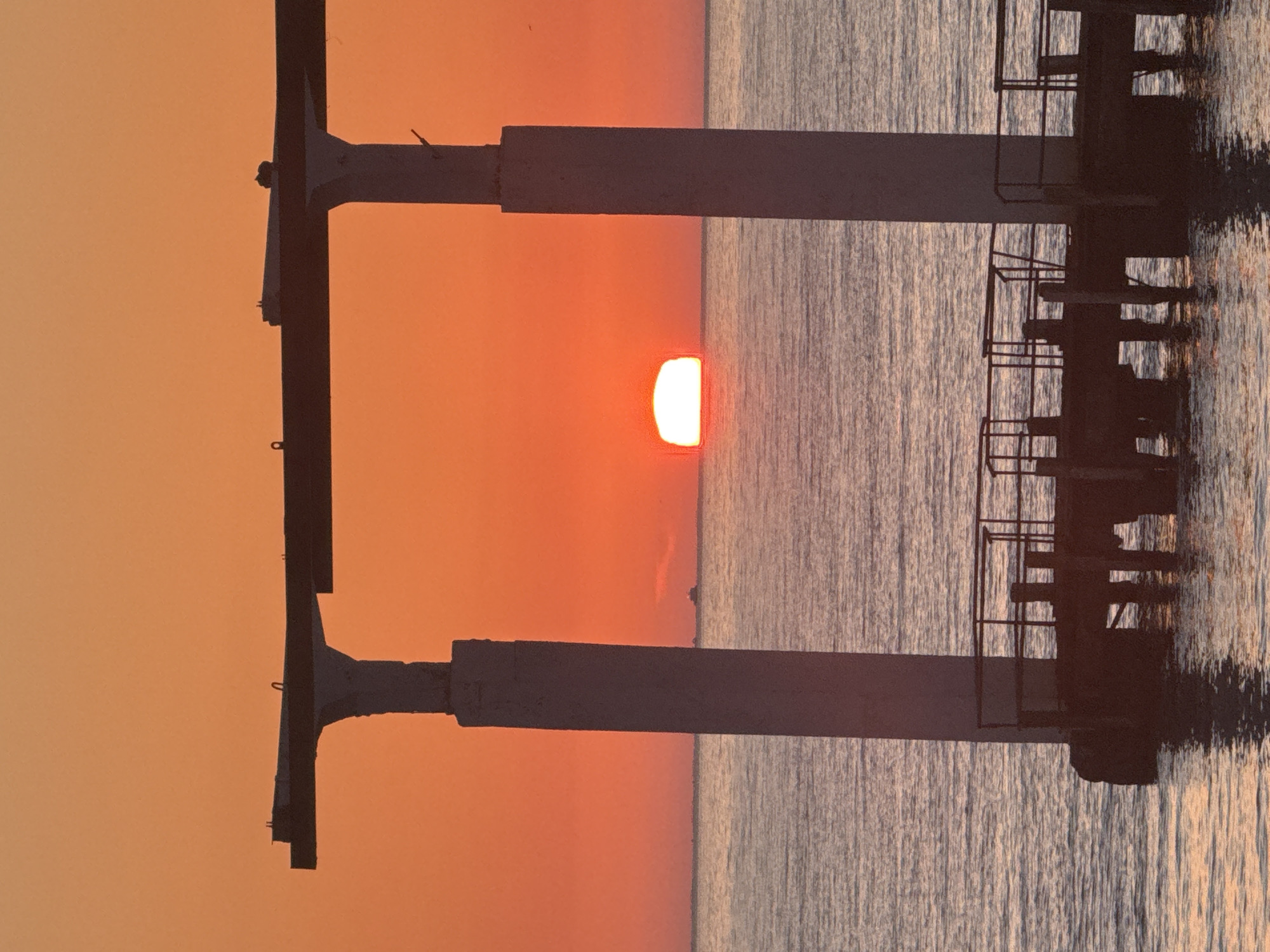 The image shows a pier or structure in the water against a sunset. The sun is partially visible, positioned between the support beams of the structure. The sky is orange, and the water surface has a subtle ripple effect. Part of the structure has ladder. The location is possibly near the city of Sochi, Russia.