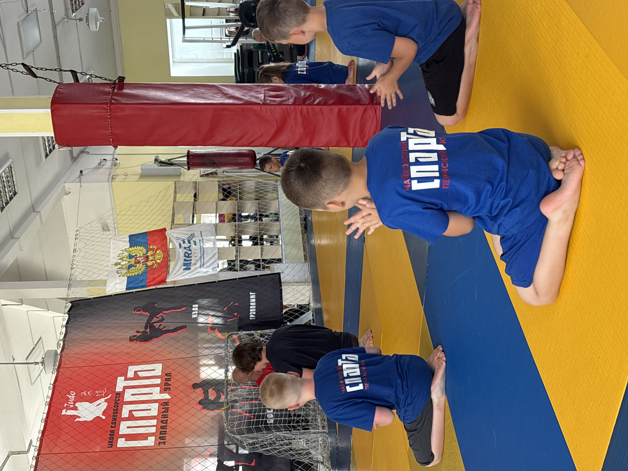 The image depicts an indoor martial arts training session, likely in Chaikovsky, Russia, given the text visible on the children's shirts. Several young boys, dressed in blue t-shirts, are sitting on a yellow and blue mat. One boy in the foreground is turned away from the camera, facing a large red padded column. He is wearing a blue t-shirt with white Cyrillic lettering that reads "Чайковский СПАРТА" (Chaikovsky SPARTA) and "ПЕРМСКИЙ КРАЙ" (Perm Krai). Another boy to his right is also turned away, wearing a similar blue t-shirt and clapping his hands. In the background, other children and an adult are visible. A large banner with "Judo" and other Cyrillic text is displayed prominently on the wall, along with a Russian flag. The environment appears to be a gymnasium with overhead lighting and sporting equipment.