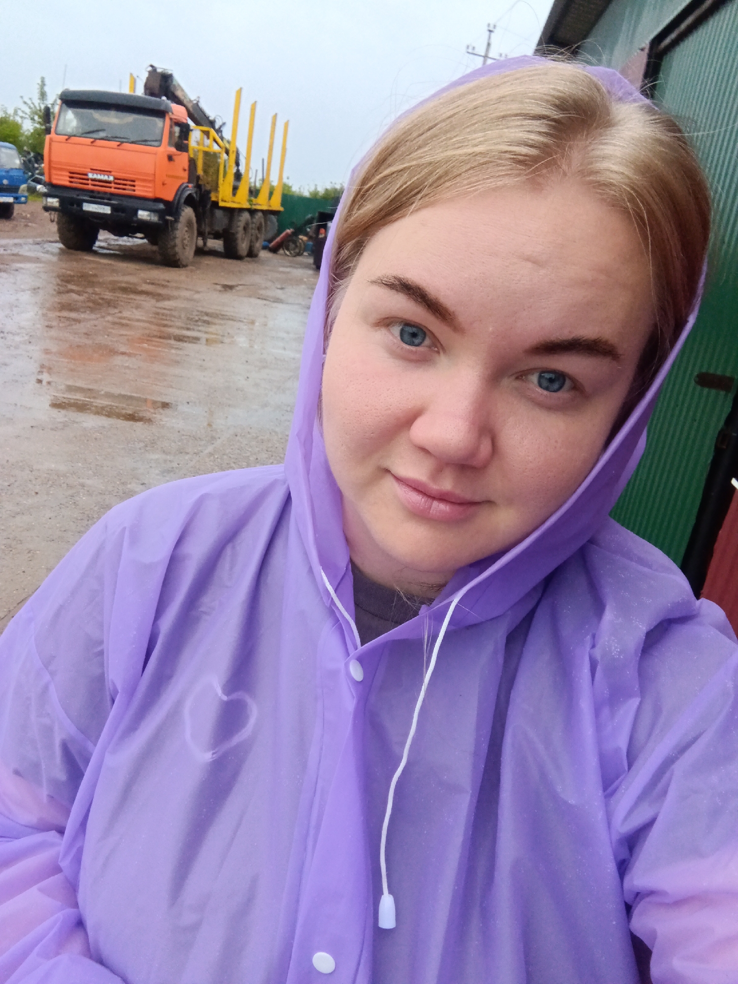 A young woman with blonde hair and blue eyes is taking a selfie in the rain. She is wearing a purple hooded raincoat with a white drawstring. A faint heart shape is visible on the chest of the raincoat. The background shows a wet, muddy ground with reflections of the sky. An orange log truck with a crane is parked to the left, its yellow logs standing upright. A blue vehicle is partially visible behind it. A green corrugated metal building is on the right. The weather appears overcast and rainy, consistent with the wet ground and the woman's raincoat. The scene suggests an outdoor industrial or logging area in Фоки, Russia. The woman has a slight smile, conveying a calm or perhaps slightly amused emotion despite the weather.