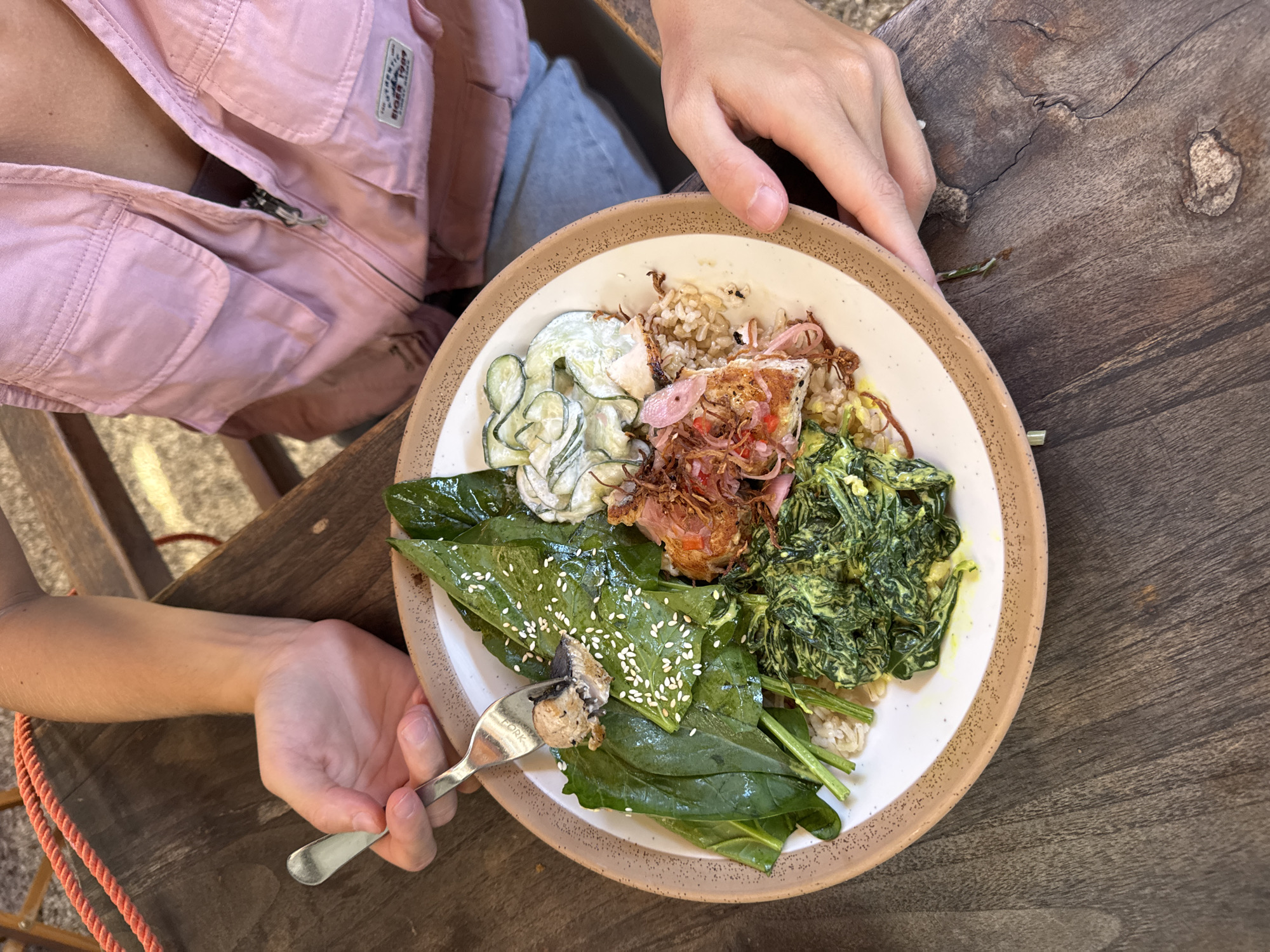 The image is a top-down shot of a plate of food being held and eaten by a person. The person is wearing a light pink jacket and light blue jeans. The plate, which is off-white with a brown rim, contains a variety of food items, including rice, cooked greens, cucumber, fried onions, and leaves sprinkled with sesame seeds. A fork is being used to pick up a piece of the food. The plate is resting on a wooden table. The photograph was taken in Kuta Selatan, Indonesia.