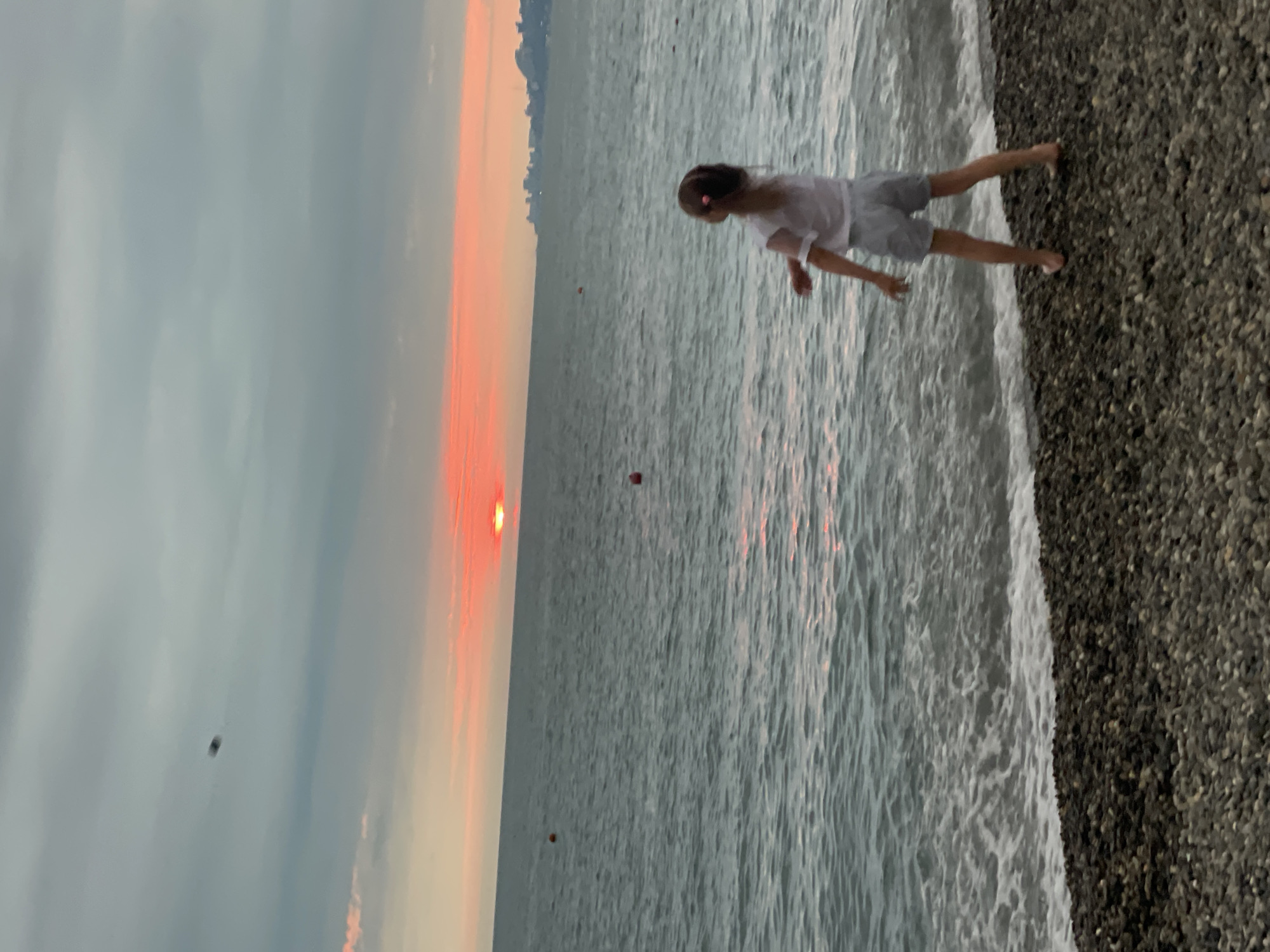 A young girl with a ponytail, wearing a white t-shirt and gray shorts, stands on a pebble beach in Sochi, Russia. She is facing away from the camera, towards a body of water where the sun is setting. The sky is cloudy, with streaks of orange and pink from the sunset. The ocean is calm, with small waves breaking on the shore. There are a few red buoys visible in the water. In the distance, a city skyline can be seen. The girl appears to be throwing something into the air, though the object is blurred.