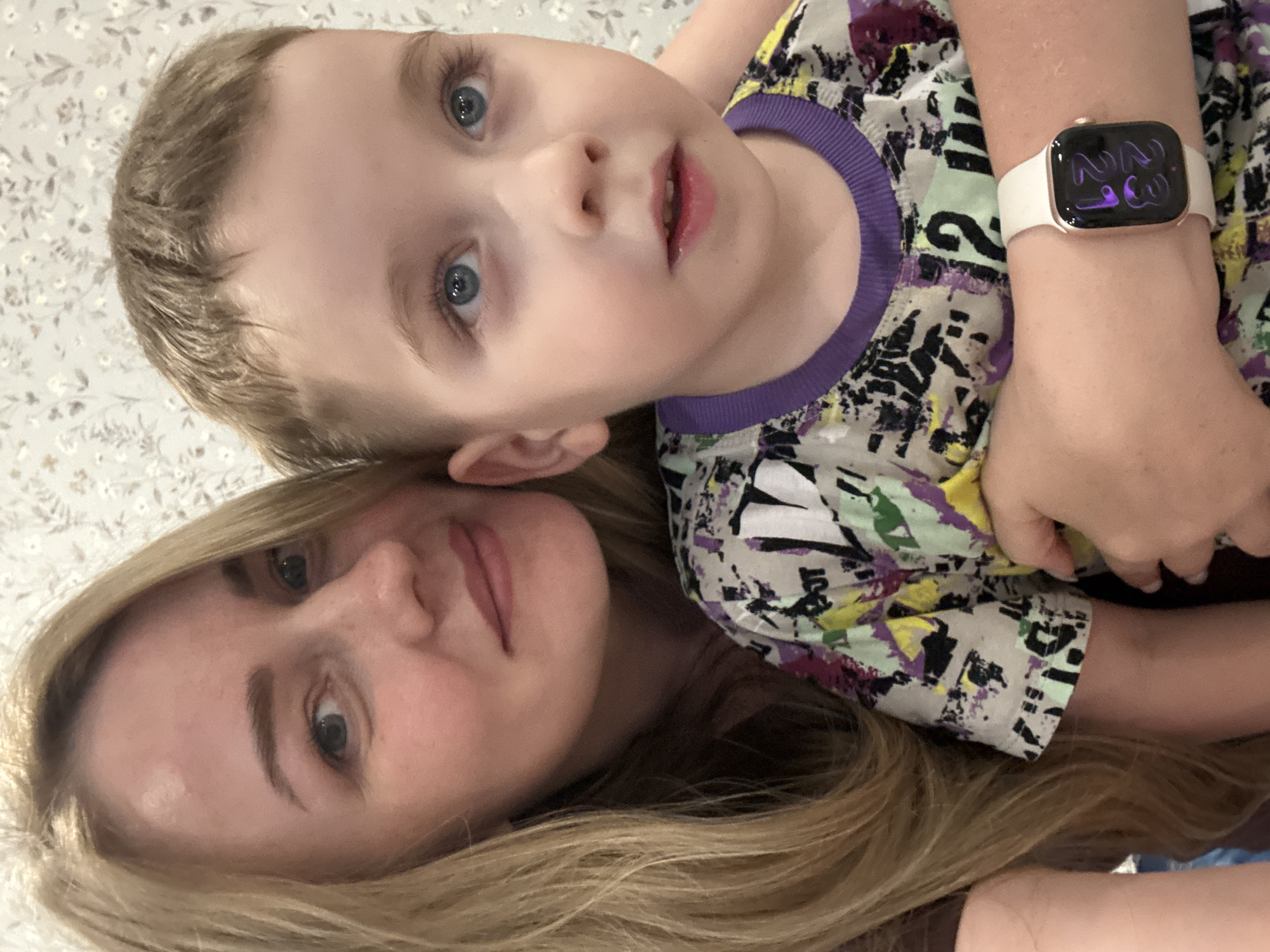 A light-skinned woman with long blonde hair is lying down facing upwards, with a light-skinned child lying on top of her. The child has blonde hair and blue eyes, and is wearing a patterned shirt with a purple collar. An adult arm wearing a watch with a white strap is visible. The background is a light colored wallpaper. The location is unknown and there is no evidence of the city of Chaikovsky, Russia.