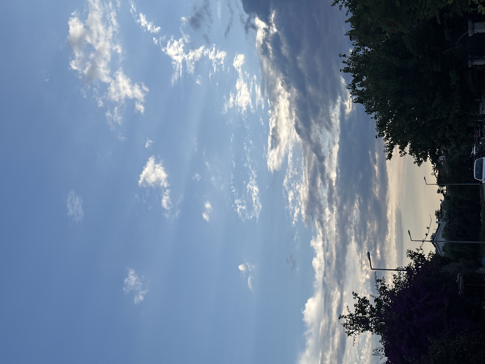 The image depicts a view in Fethiye, Turkey, during the day. A partly cloudy sky dominates the scene, with light blue sky visible amidst scattered cumulus clouds and longer streaky clouds. The sun's rays are faintly visible piercing through the clouds. The top right of the frame is partially obscured by dark green trees and foliage, indicating a roadside. Streetlights are seen protruding from the greenery. A glimpse of buildings and vehicles are visible between the trees. The image appears to be taken from ground level, looking upwards.