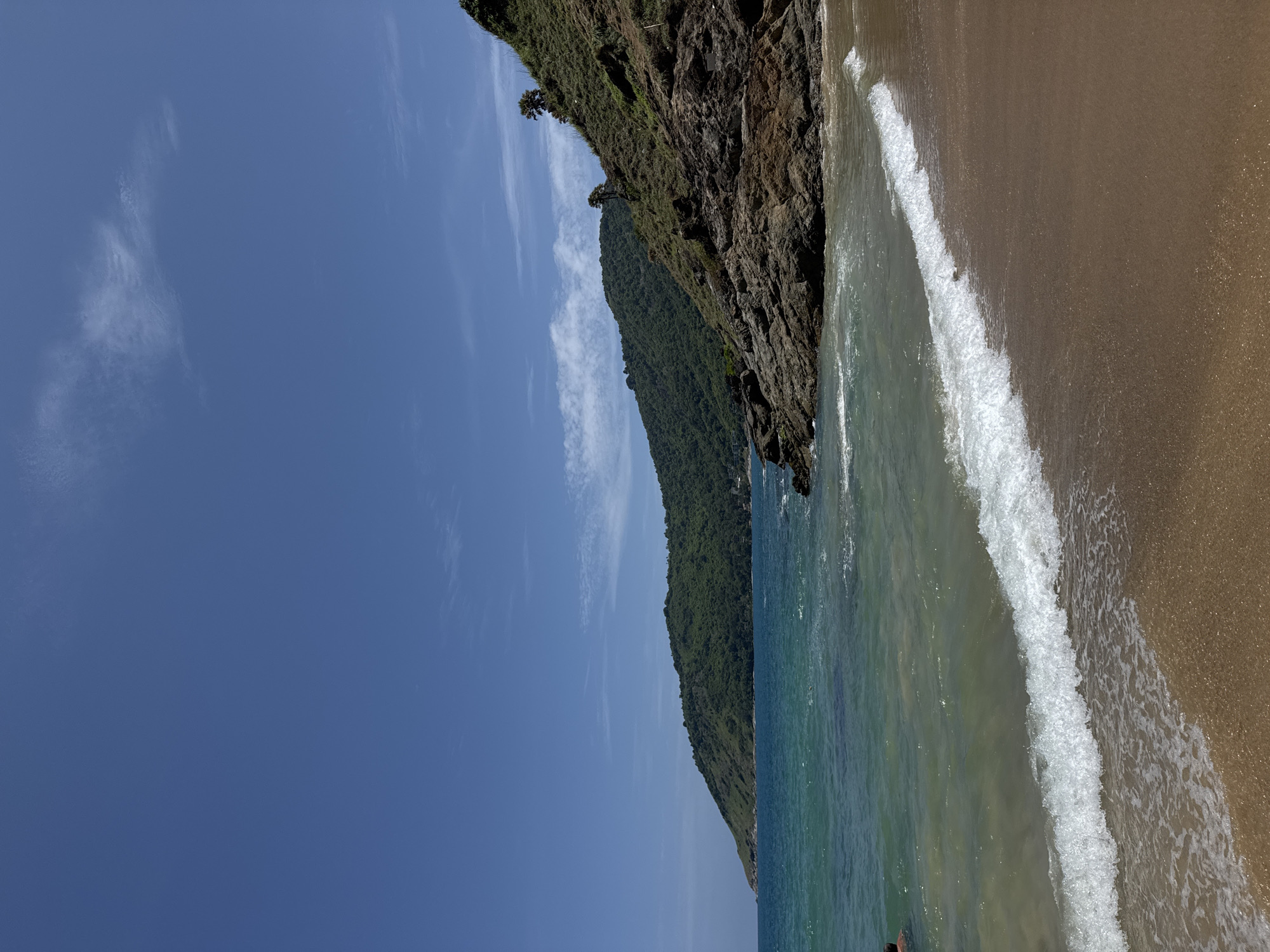 An outdoor, eye-level, full shot captures a tranquil beach scene with clear blue skies and a body of water. The image is oriented vertically. In the distance, a lush green hillside is visible. The foreground is dominated by a sandy beach with a small wave breaking at the shore. The water is a gradient of turquoise and clear, with some dark rocks jutting out into the sea on the right side of the frame. A person is partially visible swimming in the water on the left side of the image. The scene appears to be located in Rawai, Thailand.